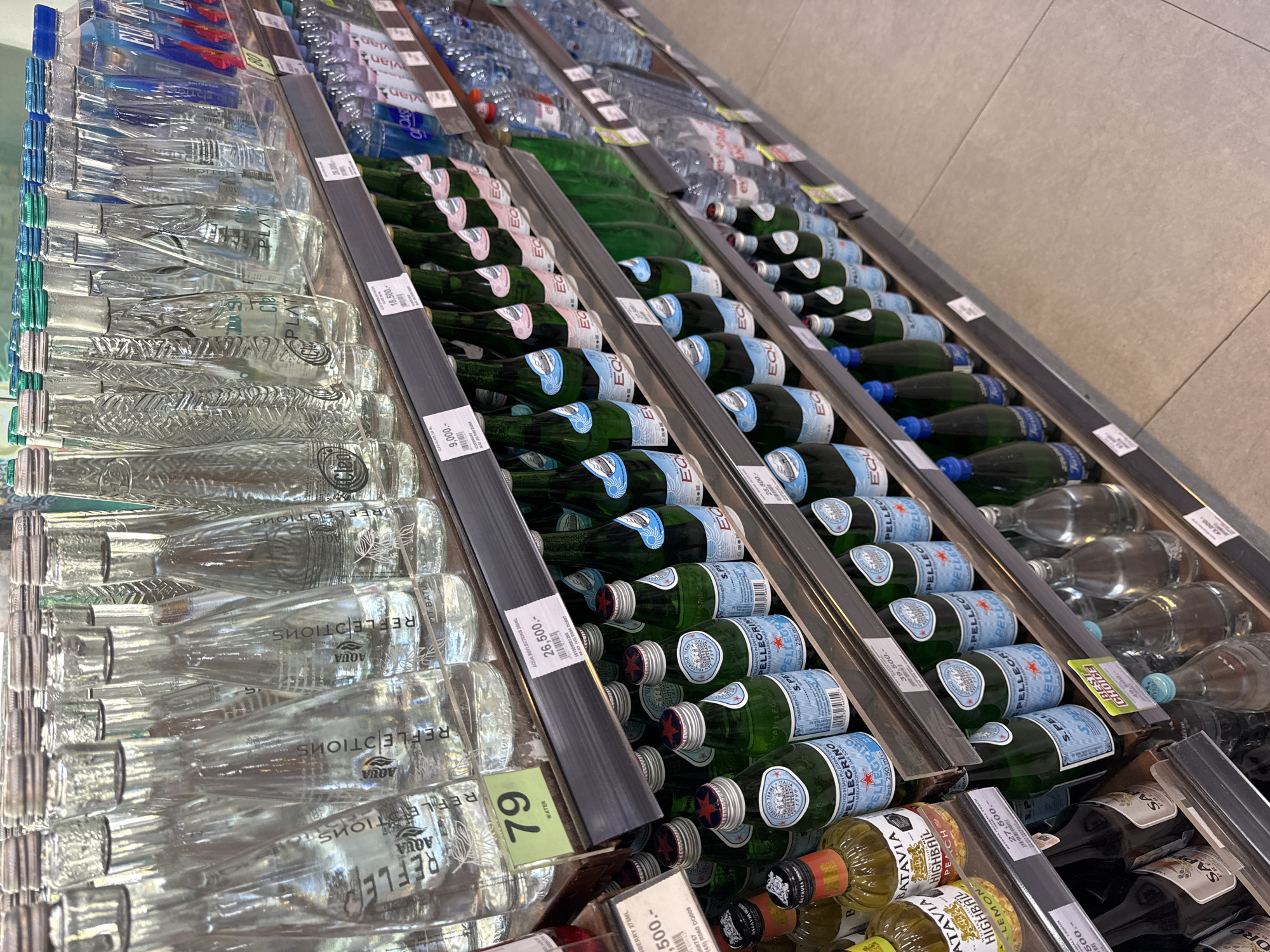This is a high-angle, slightly angled, indoor shot of shelves in a retail store, likely a supermarket or convenience store, located in Kuta Selatan, Indonesia. The shelves are stocked with various brands of bottled water and other beverages. Prominently featured are bottles of "Aqua Reflections" water on the left, rows of San Pellegrino sparkling water in green glass bottles, and also rows of Equil bottled water in between the Aqua and Pellegrino. Other brands are also visible in the background including some bottles of what appears to be Highball flavored soda (peach and lemon), and on the top shelf, the bottles are Vian and Evian water. Each shelf has a dark gray price strip that displays price tags and product information in Indonesian Rupiah. The bottles are neatly arranged in rows. The floor appears to be tiled. The lighting in the store is bright.