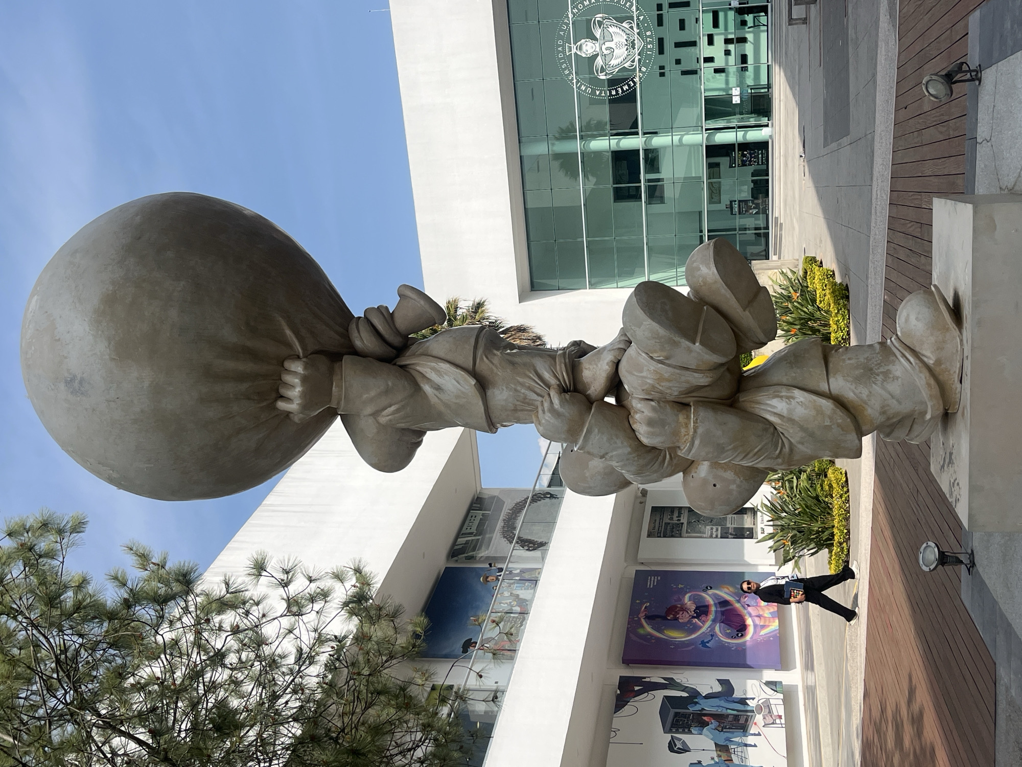 A statue depicting multiple figures stacked upon each other, carrying a large, spherical object, stands in front of a modern building with glass windows. A man wearing a black suit and sunglasses is walking along the wooden walkway next to the statue. The building has posters and artwork visible on the lower floors. A tree is partially visible on the left side of the frame. The sky is clear and blue. The location is Puebla.