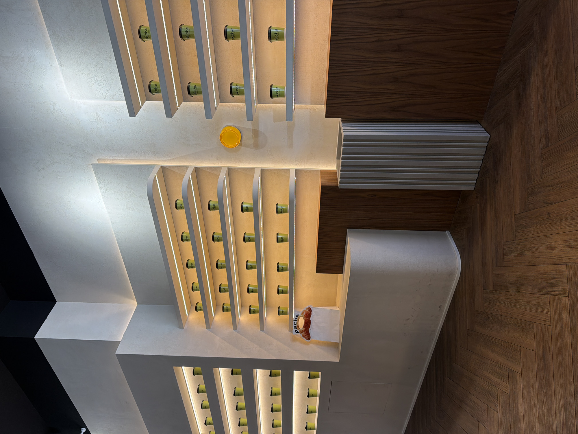 A top-down view shows a modern interior with a geometric ceiling design. The ceiling features parallel illuminated shelves containing green cups with the word "ache" written on them. A yellow lid sits near the center of the design. A bag with "pache" written on it and containing a croissant is placed on one of the white structures. The ceiling transitions into a dark wood pattern, and a corrugated metal structure is located between the white ceiling and the dark wood.