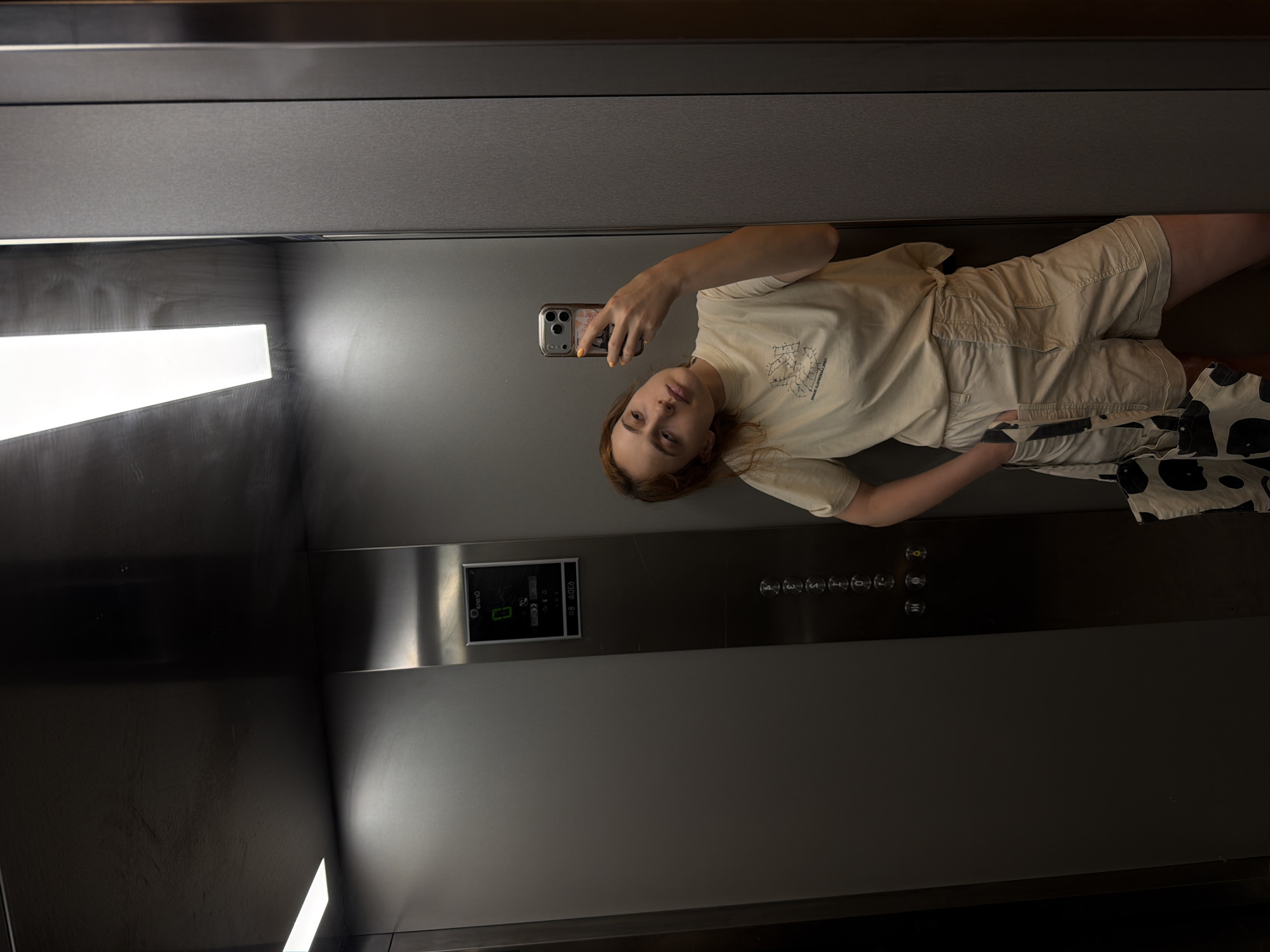 A young Caucasian woman with auburn hair and light skin is taking a selfie in an elevator. She is wearing a light beige t-shirt with a graphic design and light beige shorts. She has a black and white patterned tote bag slung over her left shoulder. The elevator has brushed metal walls and a bright fluorescent light panel on the ceiling. There is a digital display panel on the elevator wall showing the number '0'. The woman is leaning against the elevator wall with her right hand in her shorts pocket. The scene is indoors, likely in a building in Strasbourg, France.