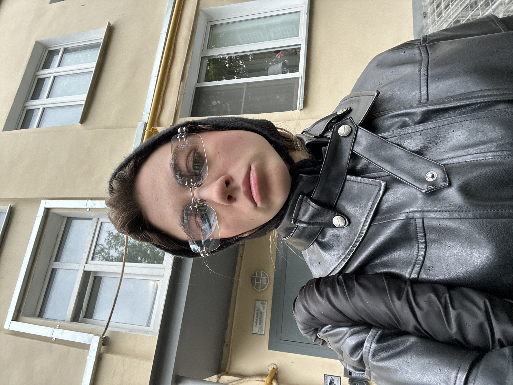 In Moscow, Russia, a young woman with dark hair, wearing silver-framed futuristic sunglasses and a black hooded jacket, stands in front of a light yellow apartment building. The building features white-framed windows. To the left, a dark gray door is visible, with a sign above it that reads "ПОДЪЕЗД № 11". A security camera is mounted near the door. The woman is holding a black, puffy sleeve to her left. The image appears to have been taken on a cloudy day, as evidenced by the overcast sky reflected in the windows.