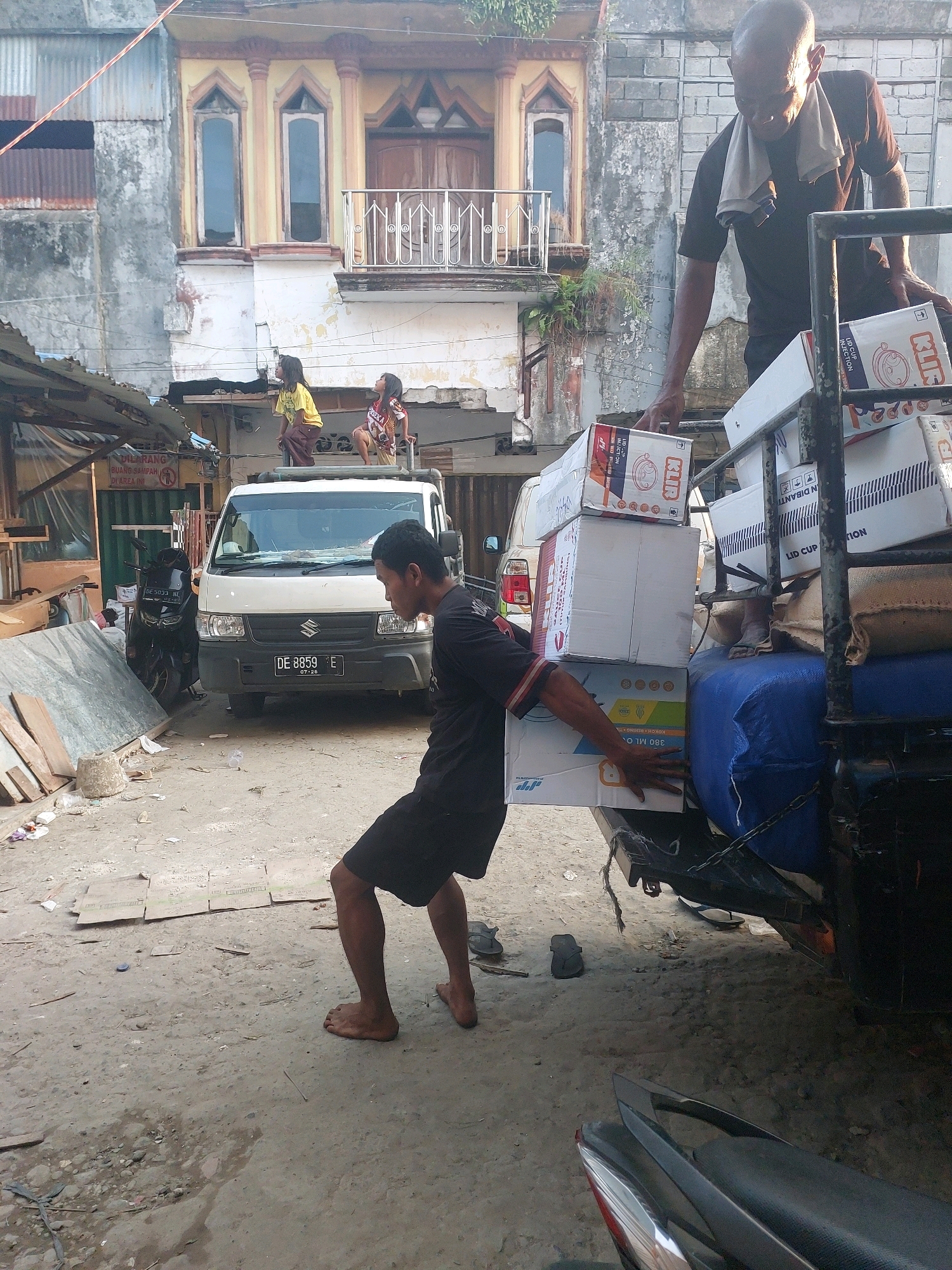 The image depicts a street scene in Ambon, Indonesia, with a man carrying a stack of cardboard boxes. He is barefoot and wearing dark shorts and a dark t-shirt, with a towel draped around his neck. The boxes are stacked precariously high and appear to be made of cardboard, with some featuring colorful labels. Behind him, a white Suzuki van is parked, and a black motorcycle is visible to the left. The environment suggests a commercial or residential area with buildings in the background, some of which show signs of age and wear. Two children are visible on a rooftop in the middle ground, looking upwards. The ground is a dirt or gravel surface, with scattered debris. The overall impression is of a busy, working-class environment.