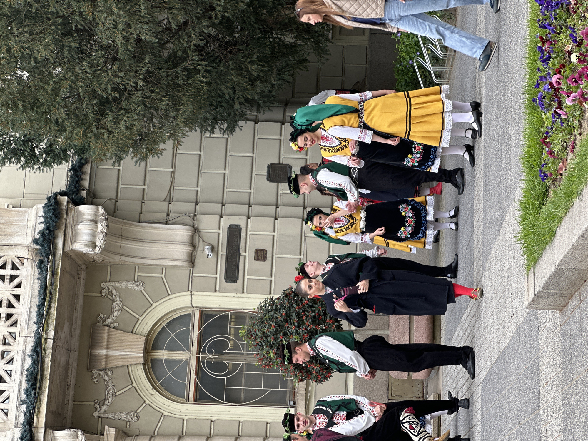 A group of people in traditional Bulgarian clothing are lined up outside of a building in Plovdiv, Bulgaria. The building appears to be a historic structure, possibly a government building. The group is a mix of men and women, and they are wearing brightly colored costumes. There is a security camera on the building to the right of the window. There are also green trees on the left and flowers on the right. A woman is walking behind them.