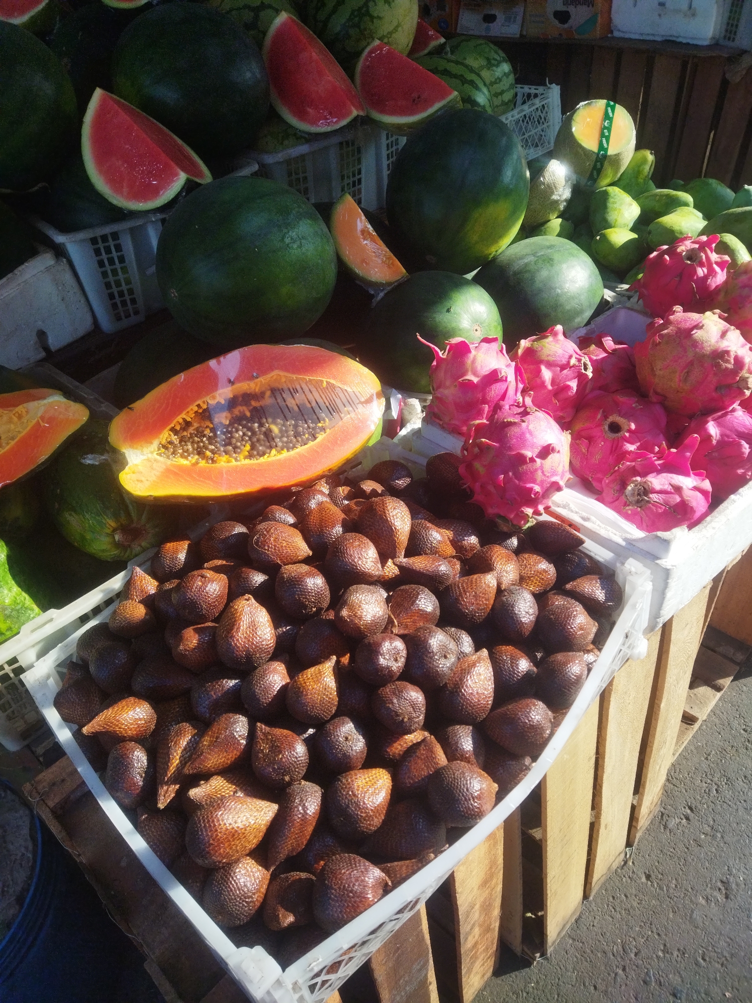 This is a high-angle, close-up shot of a fruit stand in Sukoharjo, Indonesia. The stand features a variety of fresh fruits displayed in white plastic crates and wooden crates. In the foreground, a crate is filled with salak (snake fruit), characterized by their brown, scaly skin and conical shape. Behind the salak, a sliced papaya is visible, its vibrant orange flesh and dark seeds prominently displayed. Further back, a crate holds bright pink dragon fruit, with their distinctive thorny exteriors. Several watermelons, some whole and some sliced, are also present. In the background, additional fruits like cantaloupe and various green fruits (possibly more watermelons or honeydew) can be seen. The fruits are arranged for sale, suggesting a market or roadside vendor setting. The overall impression is one of abundance and freshness, with the bright colors of the fruits contrasting against the natural tones of the crates and the asphalt ground visible at the bottom.