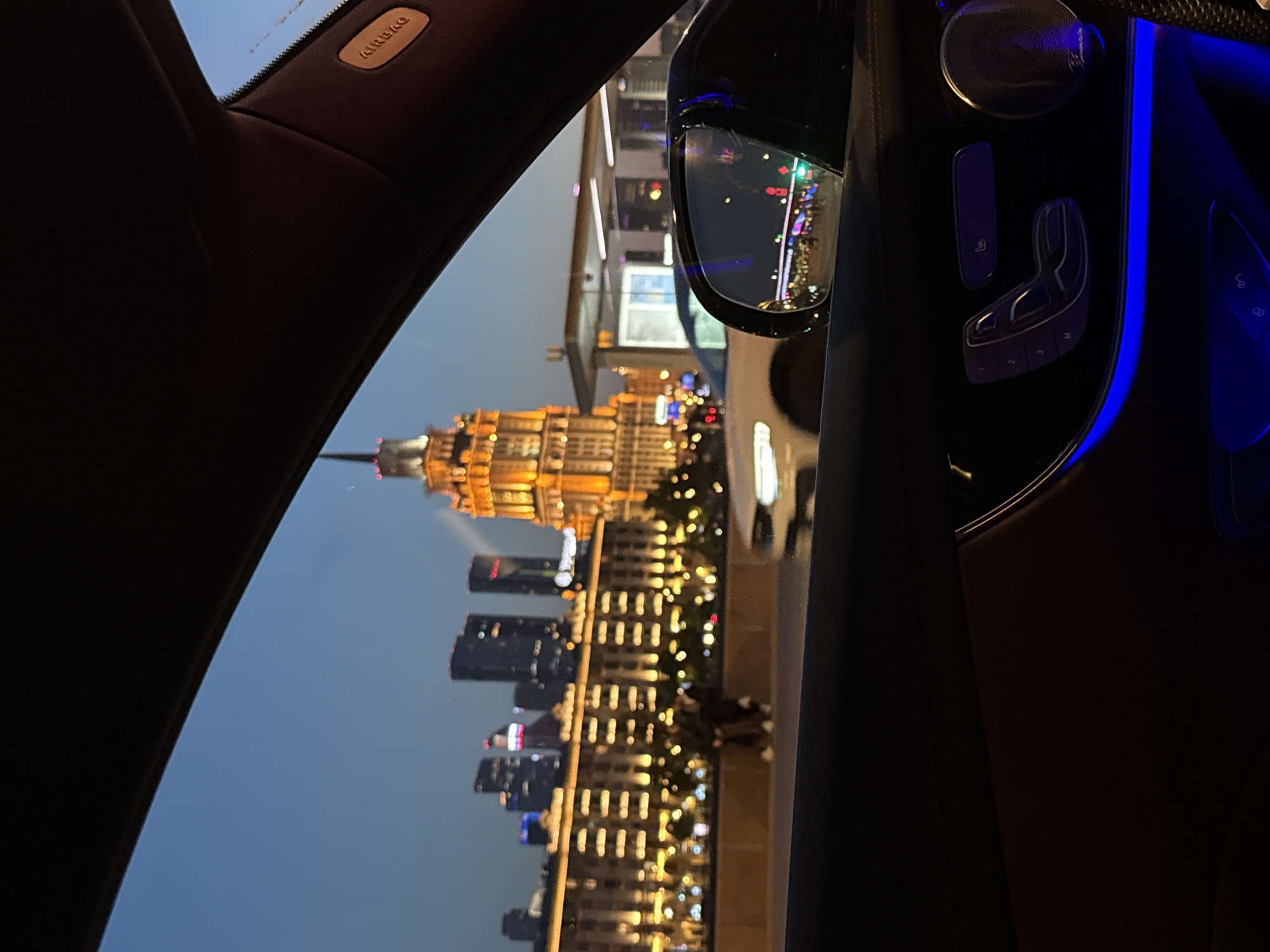 The image captures a view from inside a car at dusk, looking out through the front windshield and side window at a city skyline. The prominent structure in the background is a tall, illuminated building with a spire, characteristic of Stalinist architecture, likely one of the Seven Sisters buildings in Moscow. Several other modern high-rise buildings are also visible. In the foreground, the interior of the car is visible, including a side mirror reflecting the city lights and the car's interior ambient lighting. The side door panel with seat controls and a speaker is also visible, illuminated by blue ambient light. A white car is parked or moving on the street in front of the car from which the photo is taken. Two figures, appearing to be standing, are visible on a walkway or bridge in the middle ground. The sky is a deep blue, indicating it is either early evening or late dusk. The overall scene depicts a moment of urban nighttime travel in Moscow.