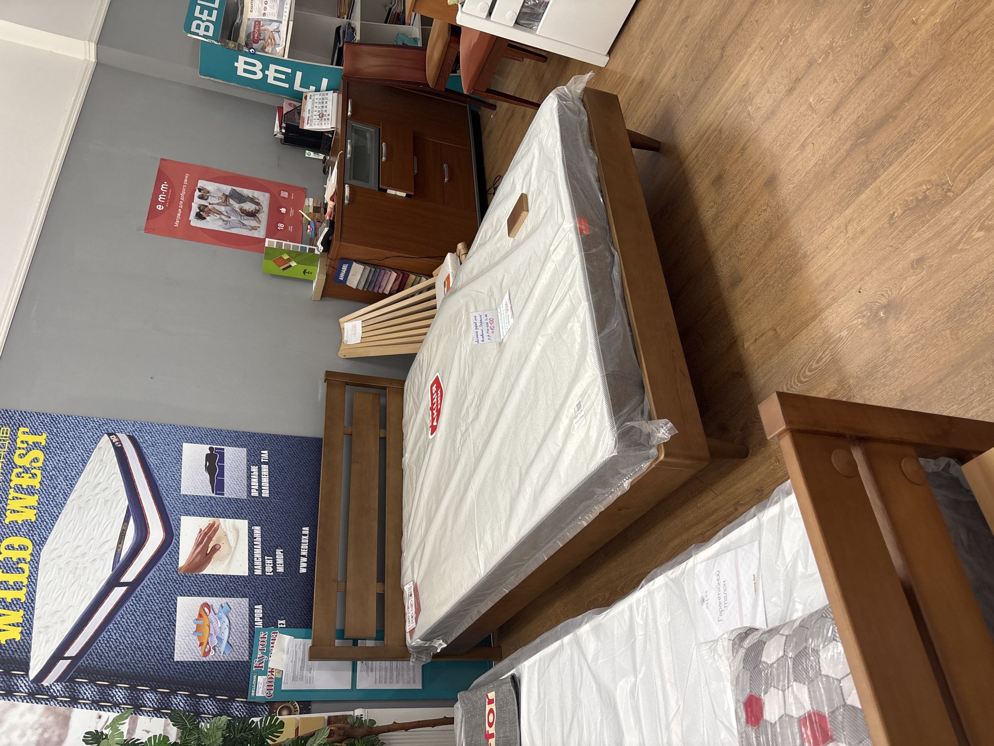 The image shows a bedroom furniture store. In the foreground, there is a wooden bed frame with a mattress covered in plastic wrap. To the left, another bed frame with a mattress covered in plastic wrap is visible. In the background, there are shelves with various products, a wooden cabinet, chairs, and posters on the wall. The store appears to be well-lit and organized.