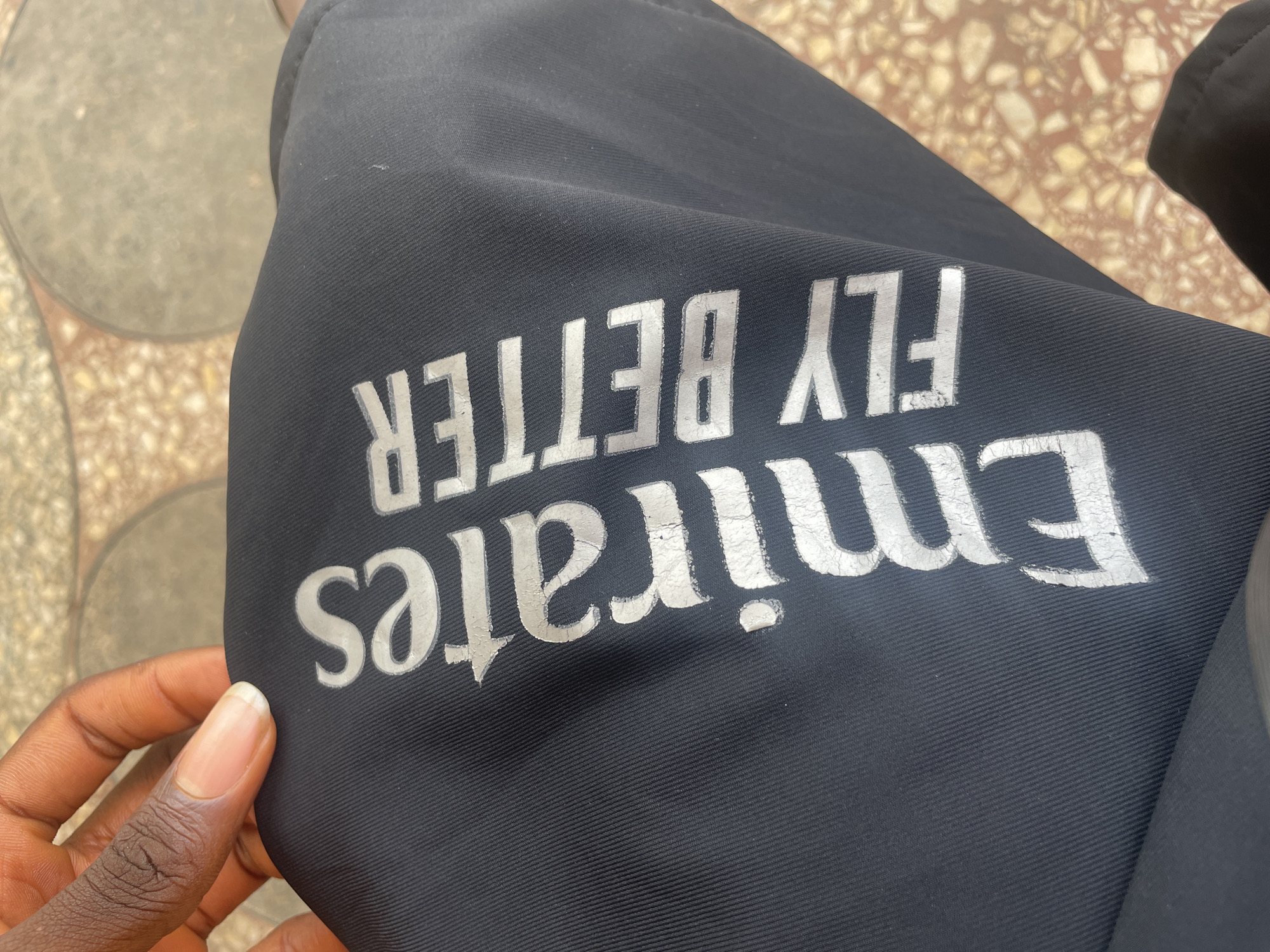 A person's hand with dark skin and a clean, light-colored fingernail is partially visible in the upper left corner of the image, touching a piece of black fabric. The fabric is adorned with the words "Emirates FLY BETTER" printed in a distressed, silver-white font. The text is oriented diagonally across the fabric, with "Emirates" positioned above and to the left of "FLY BETTER." The fabric itself has a ribbed texture. The background appears to be an outdoor setting with a patterned surface, possibly paving stones, in muted brown and beige tones. The lighting suggests it is daytime. Based on the provided information, the scene is located in Jos, Nigeria.