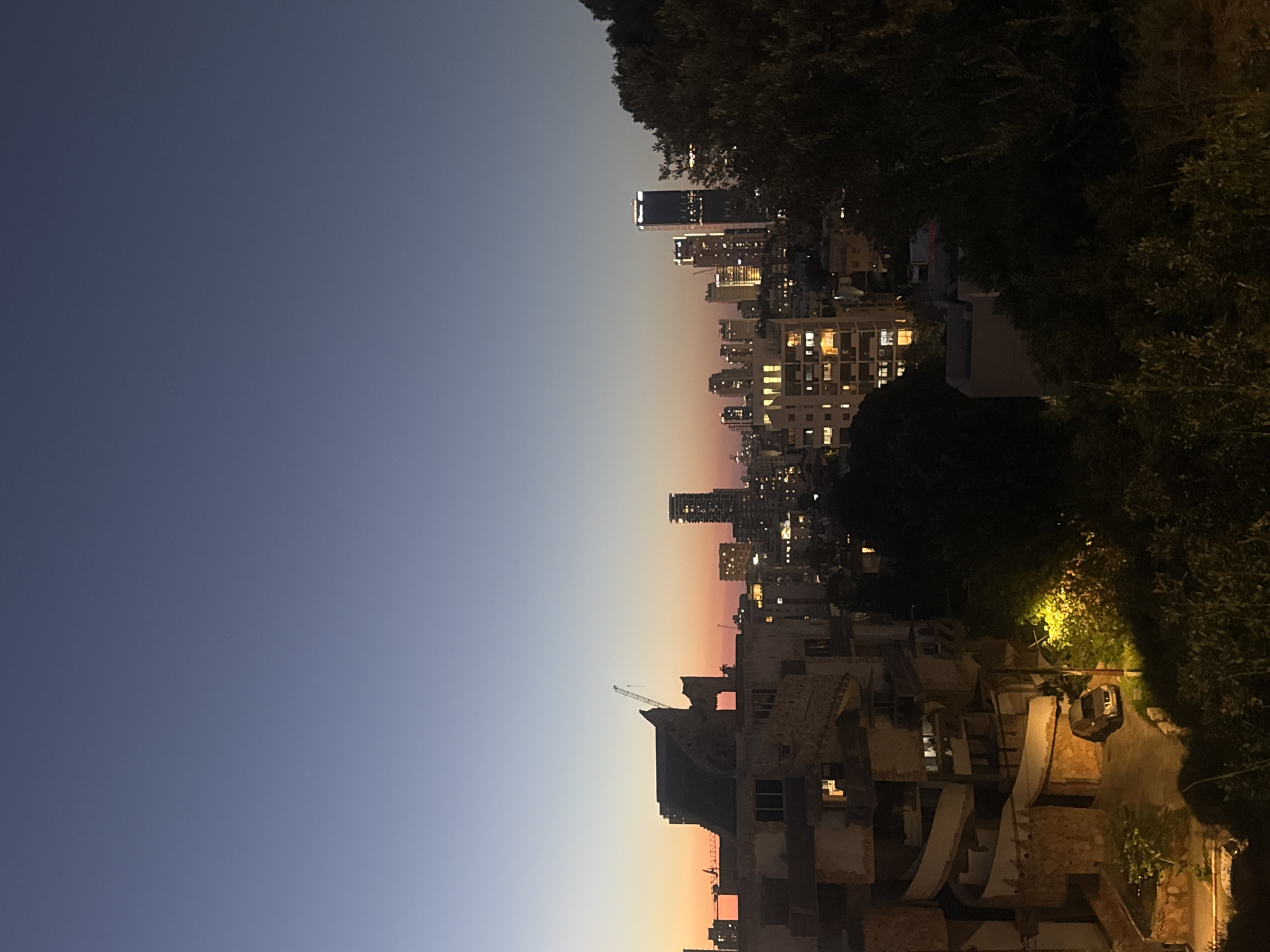 The image captures a city skyline at dusk, likely in Ramat Gan, Israel, given the distinctive architecture and the style of the illuminated buildings. In the foreground, a partially ruined, multi-story building with a Brutalist architectural style is visible. Its concrete structure shows signs of decay, with exposed rebar and incomplete sections. A spiral staircase-like element is a prominent feature of this structure. A silver SUV is parked in the paved area in front of this building, illuminated by a bright yellow streetlamp.
Beyond the foreground structure, the city unfolds with a mix of residential buildings and modern skyscrapers. Many of these buildings are lit from within, casting a warm glow against the deepening twilight sky, which transitions from a pale orange hue near the horizon to a deep blue overhead. Several construction cranes are dotted across the skyline, indicating ongoing development. Trees and dense foliage are visible in the lower right portion of the image, partially obscuring the view and adding a natural element to the urban landscape. The overall impression is of a city transitioning from day to night, with a blend of older, weathered architecture and contemporary high-rises.