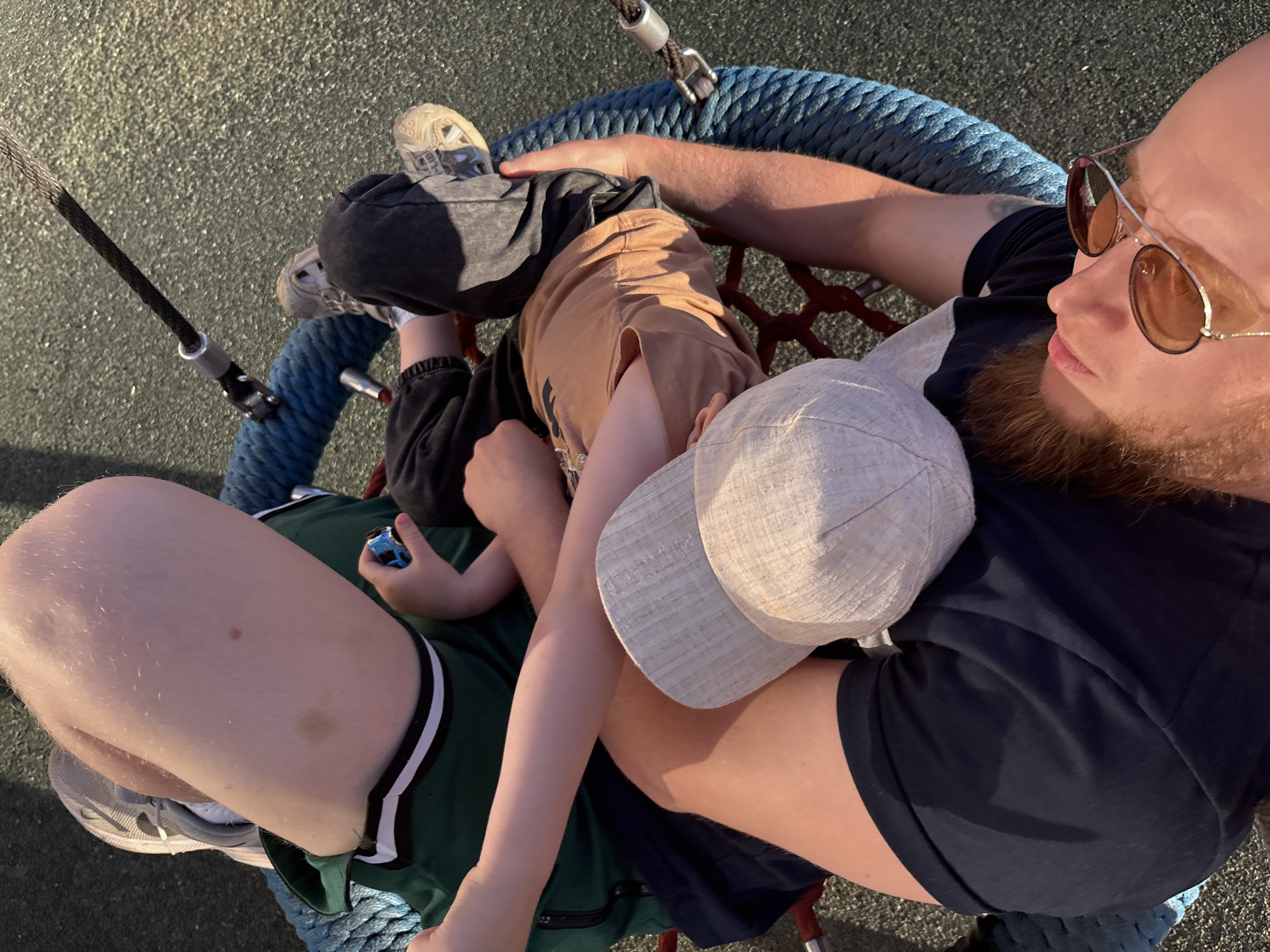 The image shows a group of people on a tire swing, likely in a park in Izhevsk, Russia. The swing is made of rope and has a circular opening.
In the foreground, a person is lying on their back, wearing a navy blue t-shirt and a light-colored baseball cap. They have a beard and are wearing sunglasses. Their arms are around another person.
To the left, another person is sitting up, wearing a green shirt with white and black trim. Their legs are extended, and they are holding a small blue toy car.
A child is visible, wearing a tan t-shirt and black pants. Their legs are bent, and their arms are resting on another person's arm.
The ground is covered with a dark, textured surface, possibly playground surfacing. The lighting suggests it is daytime, with sunlight casting shadows. The scene depicts a relaxed, familial moment.