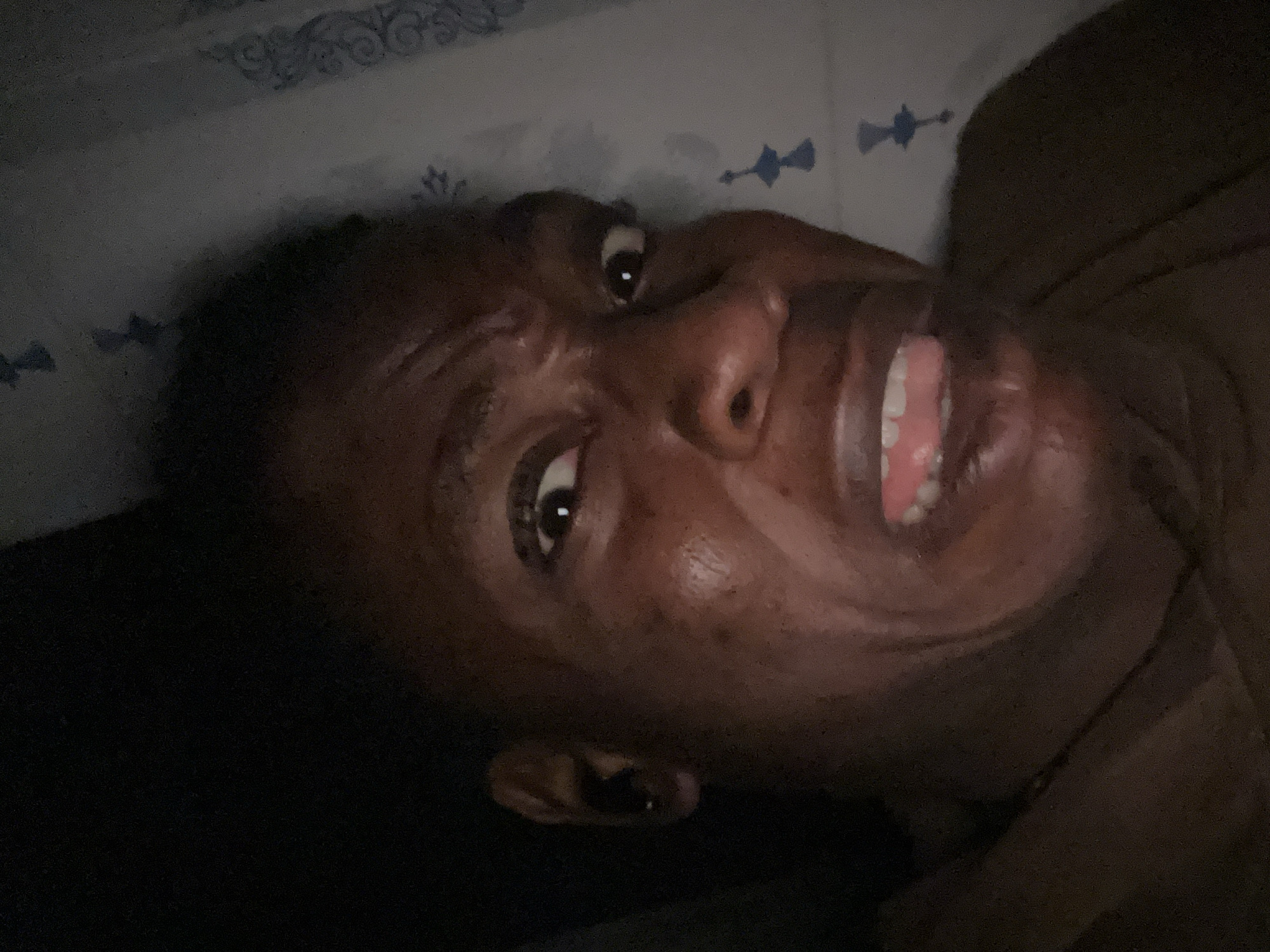 A close-up, low-angle, and partially obscured view of a dark-skinned person with their mouth slightly open revealing teeth, one ear visible with a dark-colored earring. The individual's face is angled towards the upper right of the frame. Part of a patterned pillow or sheet is visible above their head on the left, and what appears to be clothing or a blanket is visible to the right. The location is unknown.