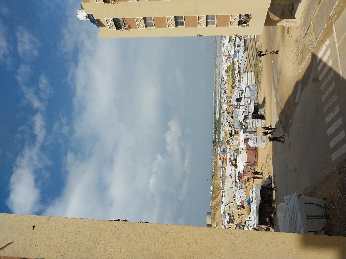 This image presents an elevated view of a densely populated temporary settlement or refugee camp in Khan Yunis, Palestinian Territory, framed by two buildings under a partly cloudy sky.
In the foreground, an asphalt road with a partial pedestrian crossing (zebra stripes) traverses the lower third of the frame, bordered by exposed dirt and sand. A substantial, angular shadow, cast by the building on the right, extends diagonally across the road and the adjacent dirt area. To the extreme left, a textured, light-brown building wall rises vertically. On the right, a multi-story, beige-colored building with brown architectural trim and multiple windows featuring decorative grilles is visible. A white water tank and blue solar panels are situated on its roof.
The middle ground is dominated by an extensive array of tightly packed, makeshift shelters and tents, indicative of a refugee camp. These structures exhibit varying materials, primarily white, grey, and blue tarpaulins, alongside some constructed from what appears to be corrugated metal or salvaged sheets, all situated on sandy terrain. Numerous individuals are present within the scene. A group of approximately five figures, predominantly dressed in dark clothing, are observed walking on the asphalt road from left to right. Additional smaller figures are visible moving along dirt paths between the shelters within the camp, with one individual in a red garment walking on a dirt path to the right of the main road.
The background shows the camp extending towards a flat, somewhat hazy horizon. On the far left horizon, a large, light-colored cylindrical structure, possibly an industrial tank or silo, is discernible atop an elevated mound. The sky above is blue, populated by scattered white and grey cumulus clouds.