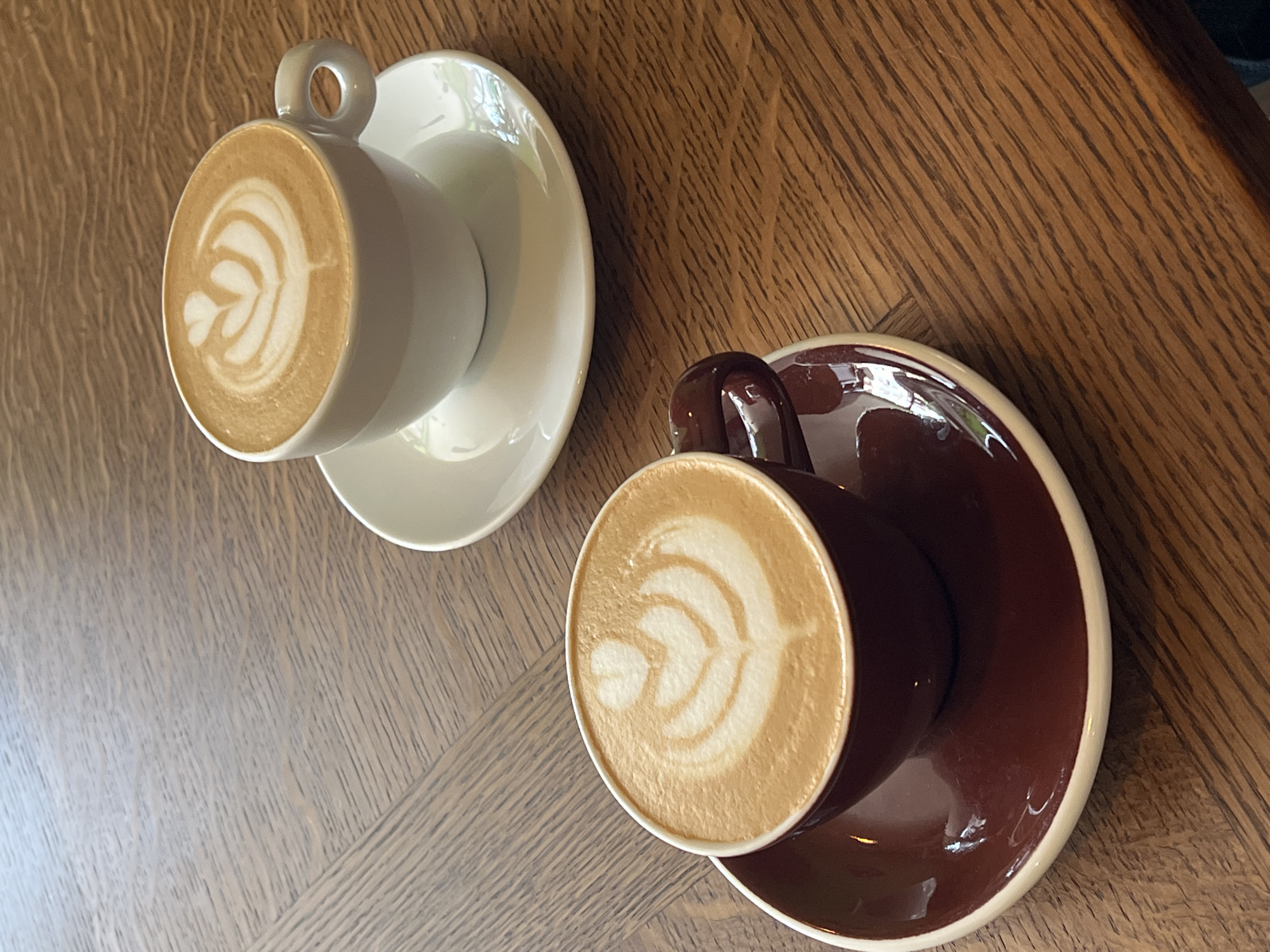 Two cups of coffee with latte art sit on a wooden table. The cup on the left is white, while the cup on the right is brown. The table is made of light-colored wood with a subtle grain pattern. The scene appears indoors. Location is unknown and can not be determined from visual data.