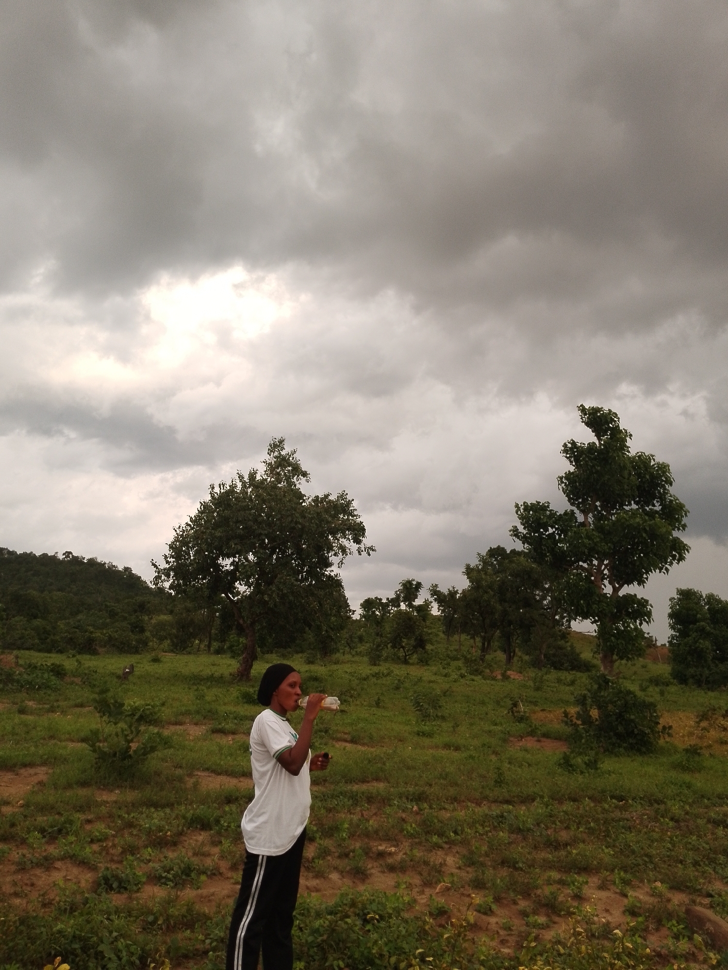 A young woman stands in a field of green grass and scattered trees, drinking from a bottle. She wears a white t-shirt, black pants with white stripes, and a black headscarf. The sky above is filled with dramatic, dark clouds. The landscape is gently rolling, with trees dotting the hills in the background. The area appears to be rural, likely in or around the city of Balewa, Nigeria, given its characteristic vegetation and atmospheric conditions. The image captures a moment of pause amidst what appears to be an impending storm.