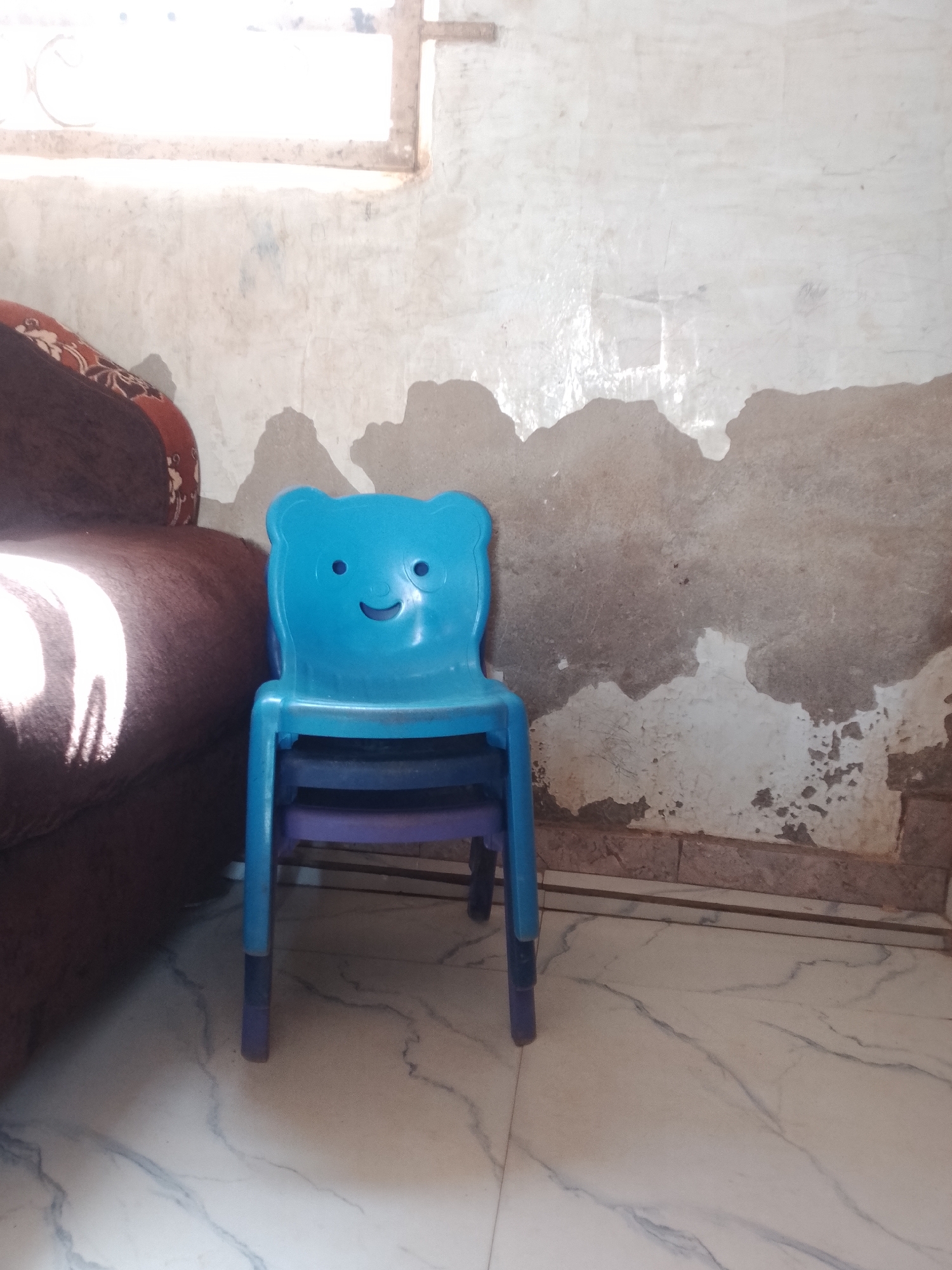 A blue, bear-shaped plastic chair is stacked on top of two other chairs, one purple and one with a dark blue seat. The chairs are positioned in front of a peeling, textured wall that is light gray with darker patches. To the left of the chairs, a portion of a brown sofa with a patterned cushion is visible. Sunlight is streaming in from an unseen window on the upper left, casting bright highlights on the sofa and creating shadows on the wall. The floor is tiled with a white and gray marble pattern. There is no visible text. The scene appears to be indoors, possibly in a child's room or play area, with a casual and somewhat worn atmosphere. The lighting suggests it might be daytime.