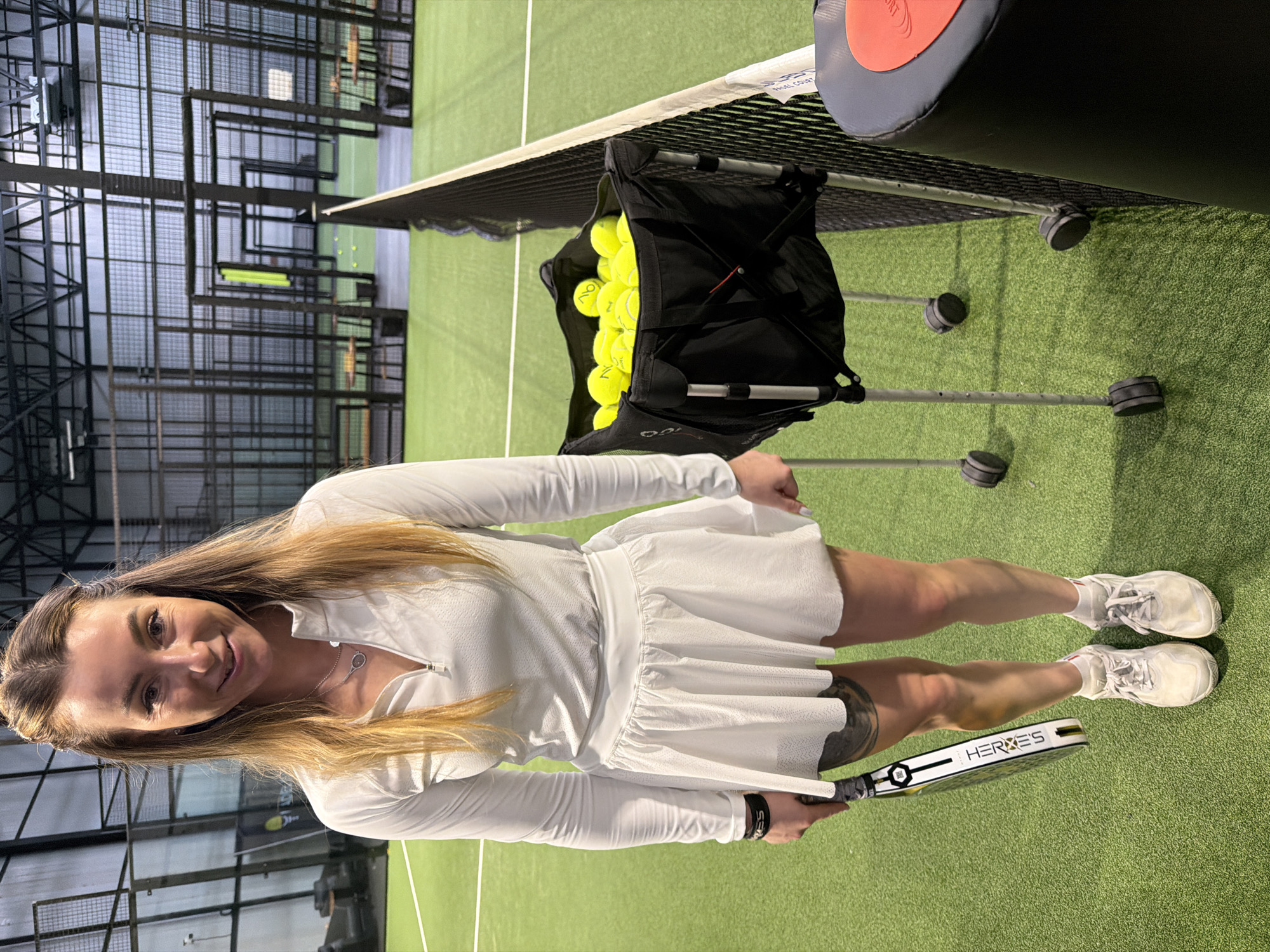 A woman stands on a green artificial turf court, possibly a tennis or padel court, in an indoor facility. She is wearing a white long-sleeved athletic top and a white pleated skirt. She holds a white and black racket in her left hand. To her right, a black wheeled basket is filled with yellow tennis balls. The background shows a netted enclosure, stadium seating, and additional courts. The environment appears to be a sports facility. The woman is smiling at the camera. There is no information in the image to suggest this location is in Choeng Thale, Thailand.