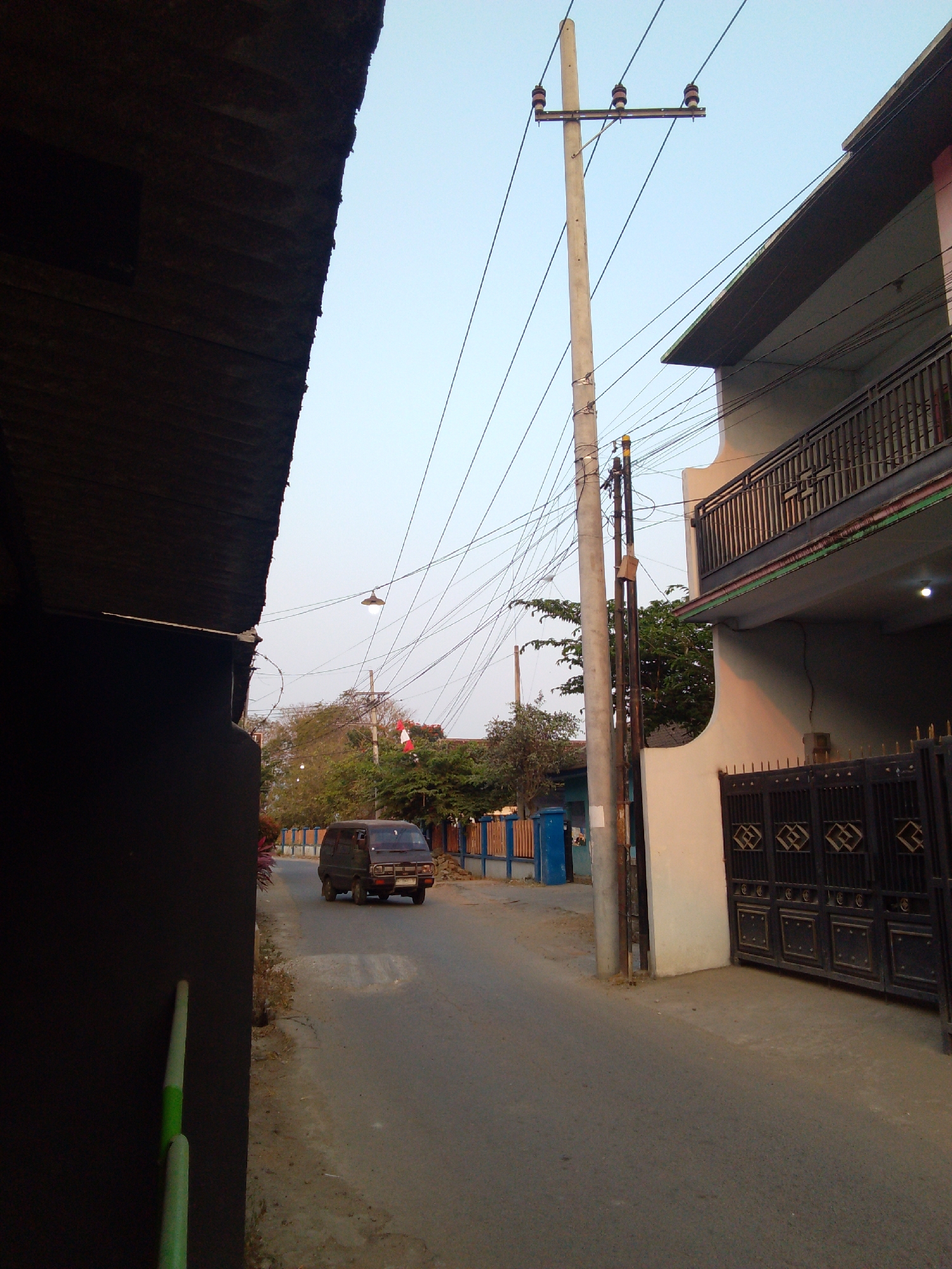 The image depicts a narrow asphalt road in a residential area of Trenggalek, Indonesia, bordered by buildings and vegetation under a clear sky. On the left, a dark-colored structure with a corrugated roof overhang partially obstructs the view. To the right, a two-story building features a balcony with a black railing and a cream-colored facade. A black metal gate with intricate geometric patterns is situated in front of this building.

A dark brown minivan is driving down the road away from the viewer. A utility pole with multiple wires stretches across the scene, supporting electrical lines. In the background, trees and foliage are visible, along with some blue fencing and a blue cylindrical trash bin. The overall scene suggests a quiet, possibly early morning or late afternoon setting in a tropical urban environment.