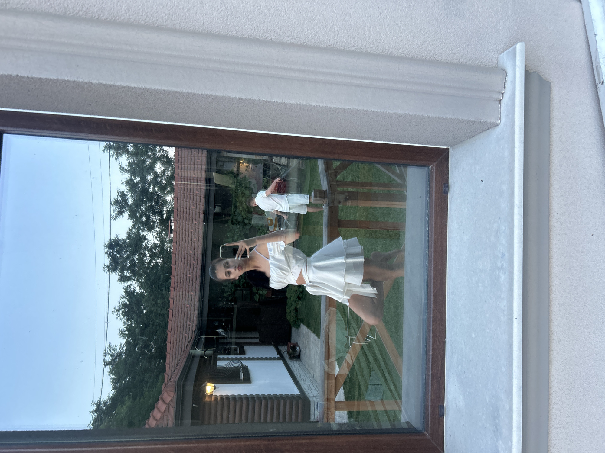 A young woman in a white two-piece outfit, consisting of a cropped top and a tiered skirt, is taking a selfie in a reflection on a window. She is standing with her left leg bent and resting on a wooden railing, while her right leg is extended. In the reflection, a man in a white t-shirt and shorts is visible watering the grass with a hose in the background. The scene appears to be a residential area, possibly a backyard, with a house with a red tile roof and greenery visible. The location is stated to be Войводино, Bulgaria.