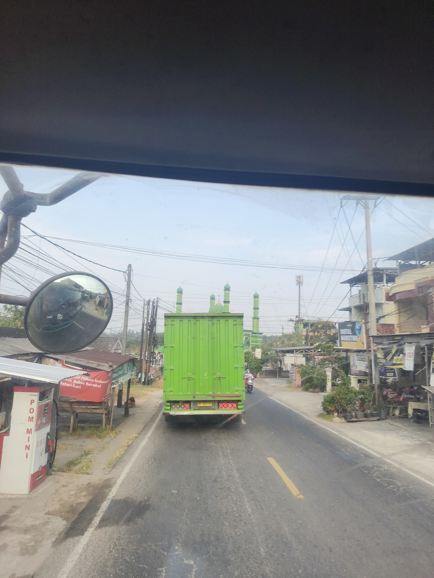 The image shows a street scene in Pangkalpinang, Indonesia. A large green truck is traveling down a paved road, which is bordered by buildings and vegetation. On the left side of the road, there is a small red and white structure with "POM MINI" written on it. Behind it, there are some small shops with red awnings. On the right side of the road, there are more buildings, some with signs that read "DIGITAL MARKETING" and "TUKANG BESI". In the distance, two green mosque domes are visible. A motorcycle is seen riding ahead of the truck. The sky is clear and bright.