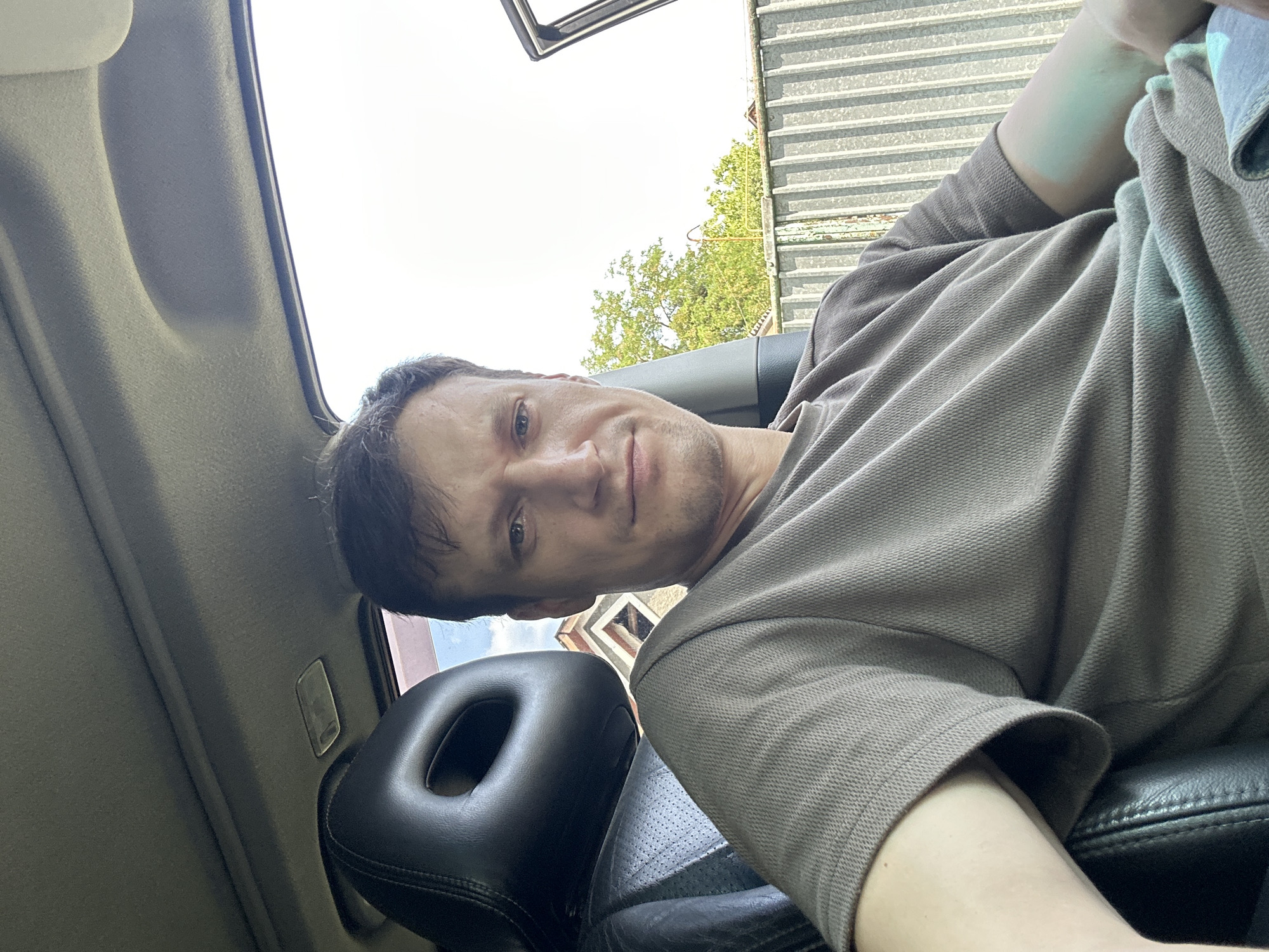 A man sits in the driver's seat of a car, looking directly at the camera. He has short brown hair and is wearing a taupe t-shirt. The interior of the car is visible, including a black leather seat and headrest. The car door is open, revealing a fence and trees outside. The man is positioned in the foreground, with the outdoor environment behind him.