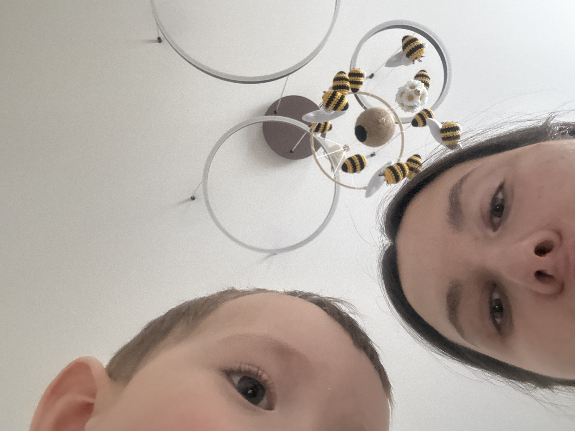 The image is a low-angle selfie or photograph taken from a point of view looking upward. Two individuals are visible, presumably a mother and a child. The adult female is positioned to the right and slightly above, while the child is to the left and slightly below. The subjects appear to be close to the camera.
Above them is a mobile hanging from the ceiling. The mobile consists of several silver rings connected to a central brown disc. Attached to the rings are knitted or crocheted bumblebee figures, a brown ball with a hole, and a white flower-like object. The ceiling in the background is plain and white.
I am unable to determine the specific location in the city of Odintsovo, Russia from this image alone.