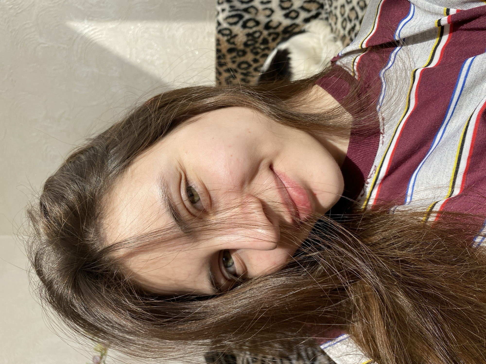 A young individual, appearing to be female, is depicted in a close-up, horizontally oriented portrait. She has long, dark brown hair, which partially obscures her face. Her eyes are open and looking towards the camera, revealing a green or hazel color. A subtle, relaxed smile is visible on her lips. She is wearing a striped top with dominant maroon, light grey, white, blue, yellow, and black stripes.
The setting appears to be indoors, likely a bedroom or living space. A leopard print fabric, possibly a blanket or pillow, is visible behind her head and shoulders. To the upper left of the frame, there's a light-colored, textured wall. Strong natural light, possibly sunlight, illuminates her face and hair from the upper left, casting distinct shadows on the wall, indicating it is daytime. The overall mood is casual and relaxed. No visible text is present in the image.