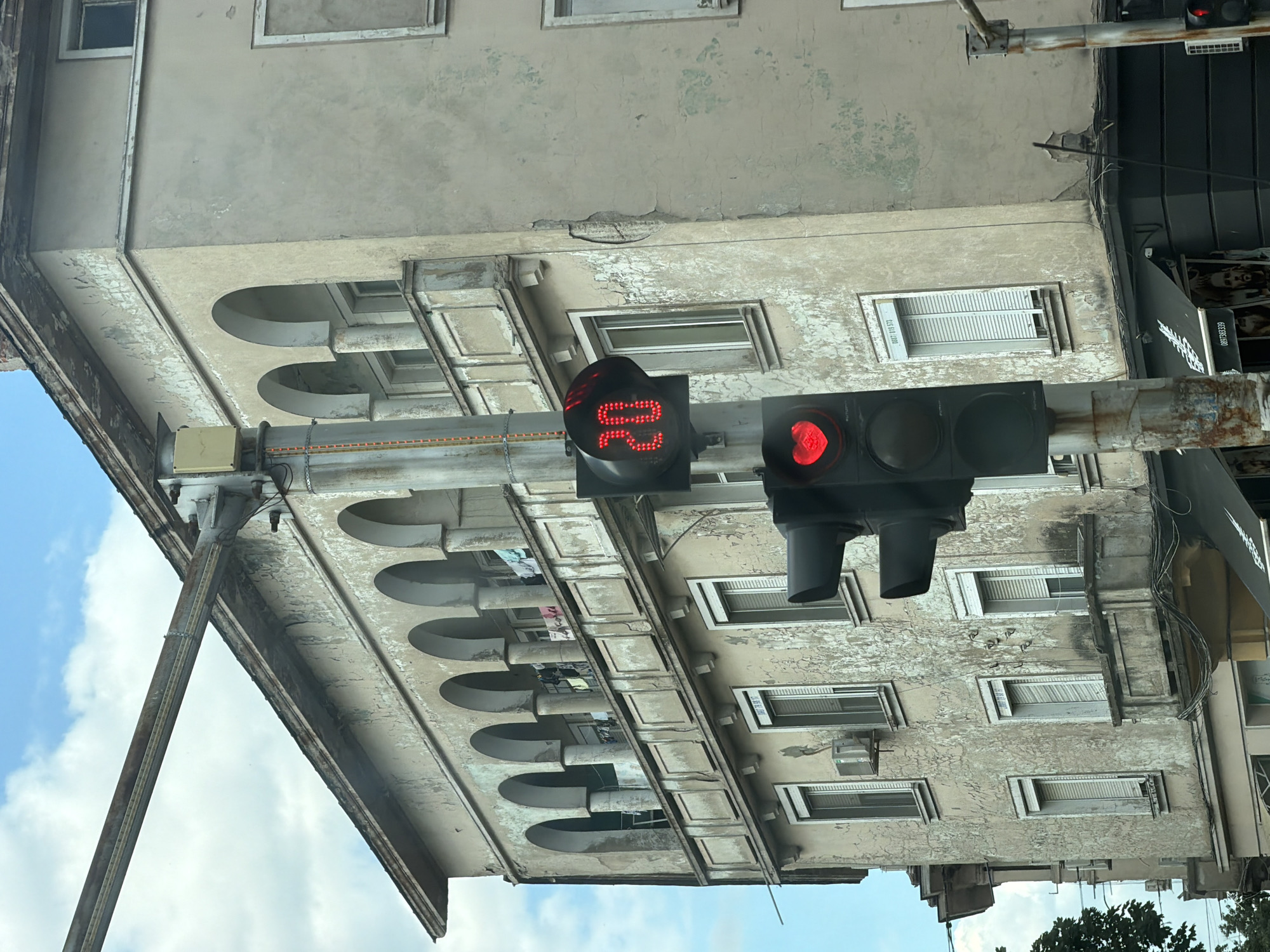In Dimitrovgrad, Bulgaria, a traffic light displays a red heart-shaped light below a red light showing the number "20." The traffic light is positioned in front of an aged, light-colored building with a weathered facade characterized by peeling paint and several balconies with arched openings. Laundry is visible hanging on a line on one of the balconies. The sky above is partly cloudy.