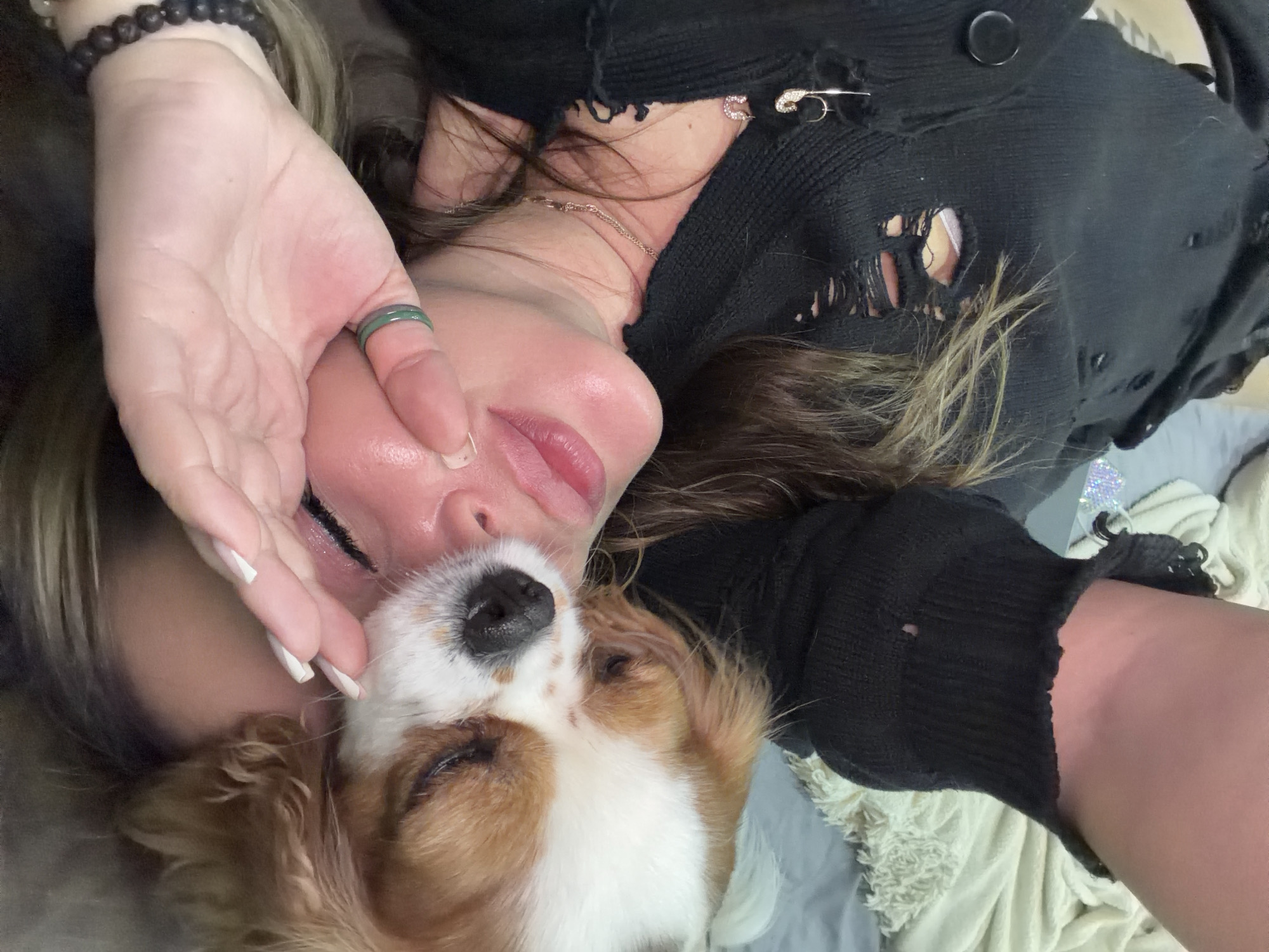 A close-up, high-angle portrait shows a person and a small dog resting together in an intimate moment of affection. The image is oriented vertically, with the subjects lying horizontally.
The human subject, appearing to be female, has long, wavy brown hair with lighter highlights. She is wearing a black, distressed knit garment, possibly a cardigan or sweater, featuring visible tears and a black button near the shoulder. Two decorative safety pins adorn her clothing: one has a sparkling "C" initial, and the other is a plain safety pin. A thin, gold-colored chain necklace is visible around her neck. Her eyes are closed or partially closed, and her lips are slightly parted, conveying a sense of deep relaxation or sleep. A hand, with white-tipped manicured nails, gently touches the dog's face. A dark, beaded bracelet is on her wrist, and a green band-style ring is on her thumb.
The canine subject is a small dog with white and light brown (tan/sable) fur, resembling a Cavalier King Charles Spaniel or similar breed. It is snuggled close to the person's face, with its eyes closed and black nose prominent, also appearing to be resting peacefully.
The background consists of soft, light-colored, textured fabric, likely blankets or bedding, indicating an indoor setting, possibly a bed or sofa. The overall ambiance is one of comfort, peace, and companionship. The image captures a quiet, tender moment between a pet and its owner. While the location is provided as Chaikovsky, Russia, there are no specific visual cues within the image itself to confirm this.