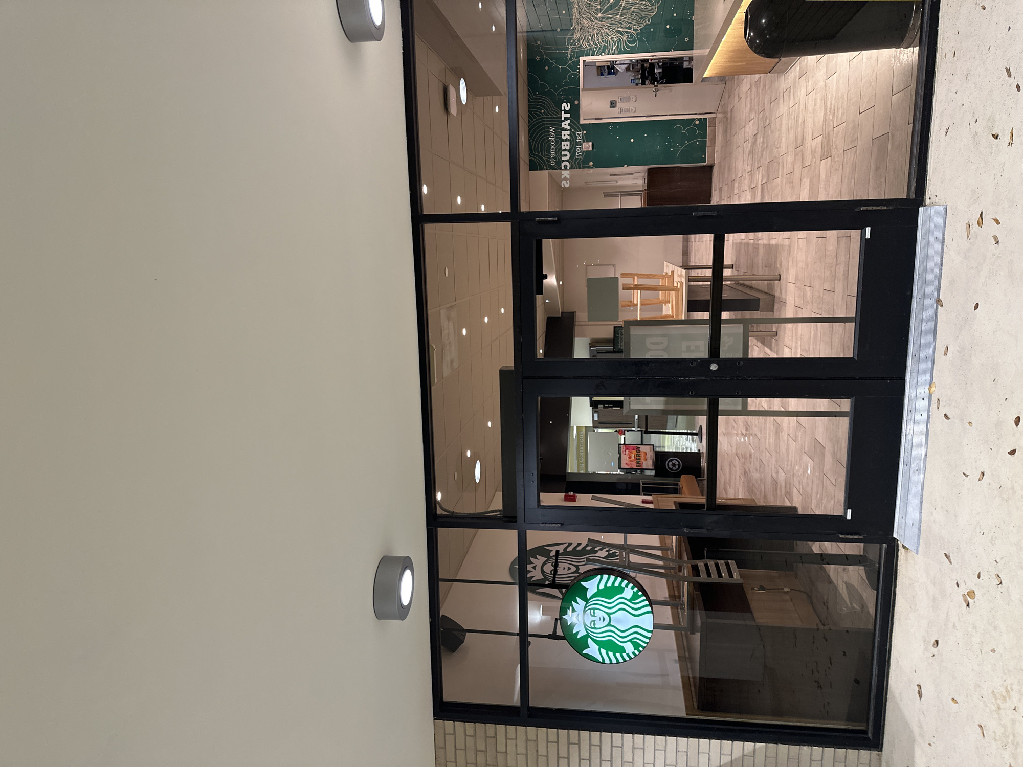 Here is a description of the image:
The image shows the entrance to a Starbucks store. The storefront features glass doors with a Starbucks logo visible on the glass. The exterior walls are light-colored and have cylindrical lights mounted on them. The walkway leading into the store is paved with light-colored tiles. The photo is taken from an external, slightly elevated perspective.
While I can describe the scene, I cannot verify that the location is Tampa, United States of America.