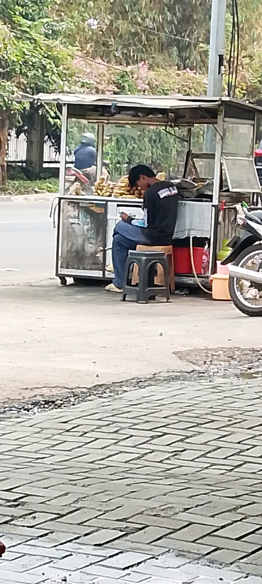 A man sits on a small stool in front of a food cart in Bekasi, Indonesia, looking at his phone. The cart has a glass display case and shelves holding food items, possibly fried snacks, and a red and orange container. A motorcycle is parked to the right of the cart. The ground in front of the cart is paved with bricks, and the area behind it is a street with trees and a utility pole. Another person wearing a helmet is visible inside the cart.