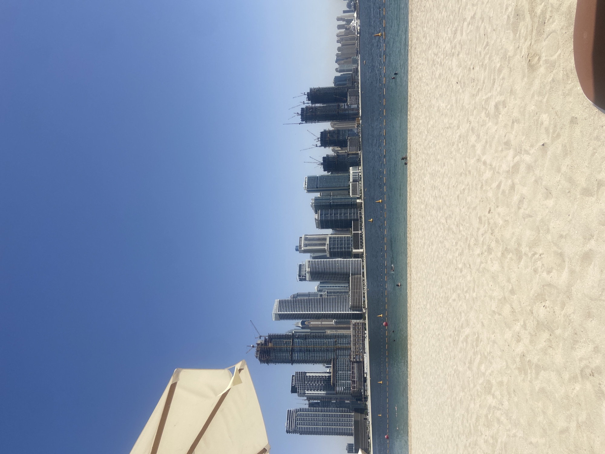 The image portrays a beach scene with the Dubai skyline in the background. The foreground features light-colored sand, and the shoreline is marked with buoys. The Dubai skyline consists of numerous high-rise buildings, some of which appear to be under construction with cranes visible. The sky is clear blue, and a beach umbrella is visible in the lower left corner of the frame. The image is rotated, presenting a vertical view of the horizontal scene. There are some people visible in the water.