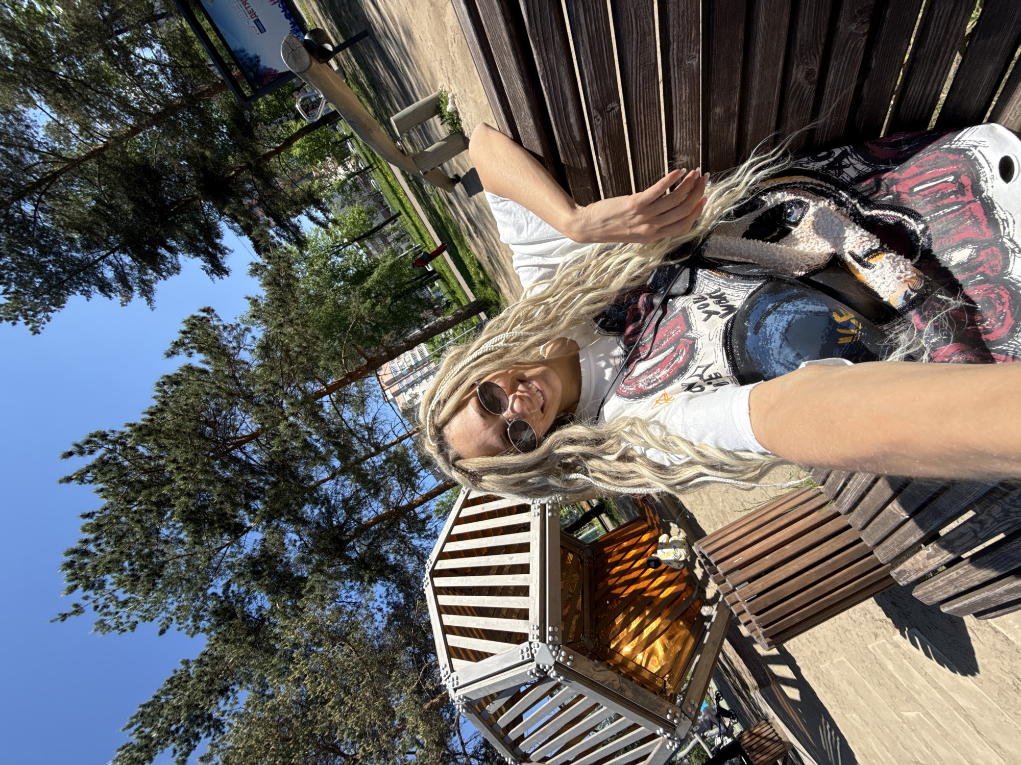 The photograph depicts a woman with long blonde hair lying on a wooden bench in a park. She is wearing sunglasses and a white t-shirt with graphics on it. A patterned bag rests beside her. The background includes trees, a blue sky, and what appears to be a modern art installation constructed of wood and reflective material. The location is a park in an unidentified city.