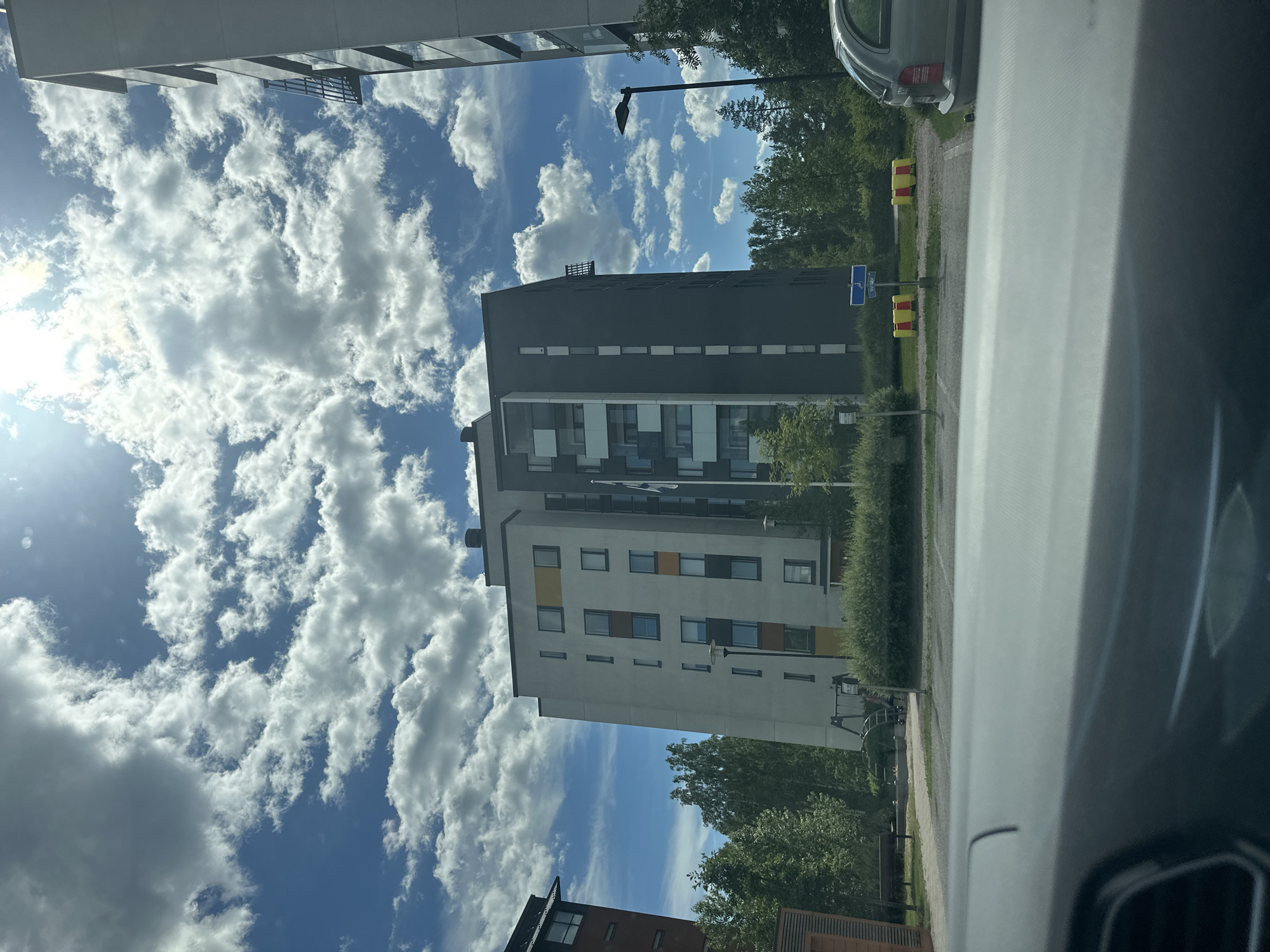 This media file is a photograph taken from inside a vehicle, looking out at an urban residential scene on a sunny day. The perspective is skewed, with the buildings and sky appearing tilted.
The primary subjects are modern apartment buildings, characterized by their clean lines and multi-story construction. One building is predominantly dark gray with white horizontal banding and rectangular windows. Another building visible has a lighter facade with colorful rectangular accents in yellow and orange, interspersed with windows. Green trees and manicured hedges surround the buildings, suggesting a well-maintained residential area.
In the background, a clear blue sky is dotted with fluffy white clouds, indicating pleasant weather. The sun is visible, casting a bright light that creates some lens flare. Elements of urban infrastructure are also present, including a street lamp and blue street signs with white text. One sign appears to be a parking sign with a wheelchair symbol. In the foreground, the interior of a car is visible, with part of the dashboard and windshield framing the outdoor scene. A light-colored car is parked in the middle ground.
There is no discernible activity or event taking place, and the scene appears to be a static view of the environment. The overall mood is one of calm and everyday life in a modern residential setting. The time of day is likely midday, given the bright sunlight. The location is urban but with a strong presence of green spaces.
No specific text that can be clearly read is visible on the signs beyond the parking symbol. The image captures a slice of life in a contemporary city.