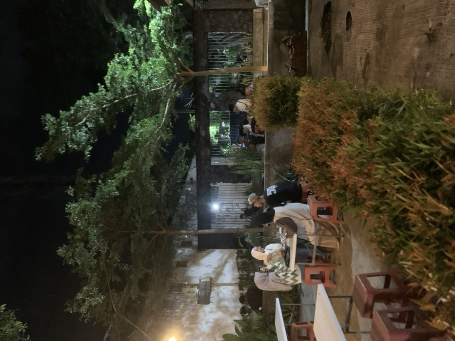The image shows a group of people sitting at a table in an outdoor seating area at night in Probolinggo, Indonesia. The location is identified by the latitude -7.751 and longitude 113.213. The scene is lit by streetlights. There are four people seated at a table with a white tablecloth. Three of them are women, wearing head coverings, and one is a man. The women are in conversation. Another man stands behind the table. There are other people seated in the background behind a low wall. The ground is concrete and covered in a layer of dried leaves. The people are sitting in a space surrounded by a hedge of plants. The scene is illuminated by overhead lighting.