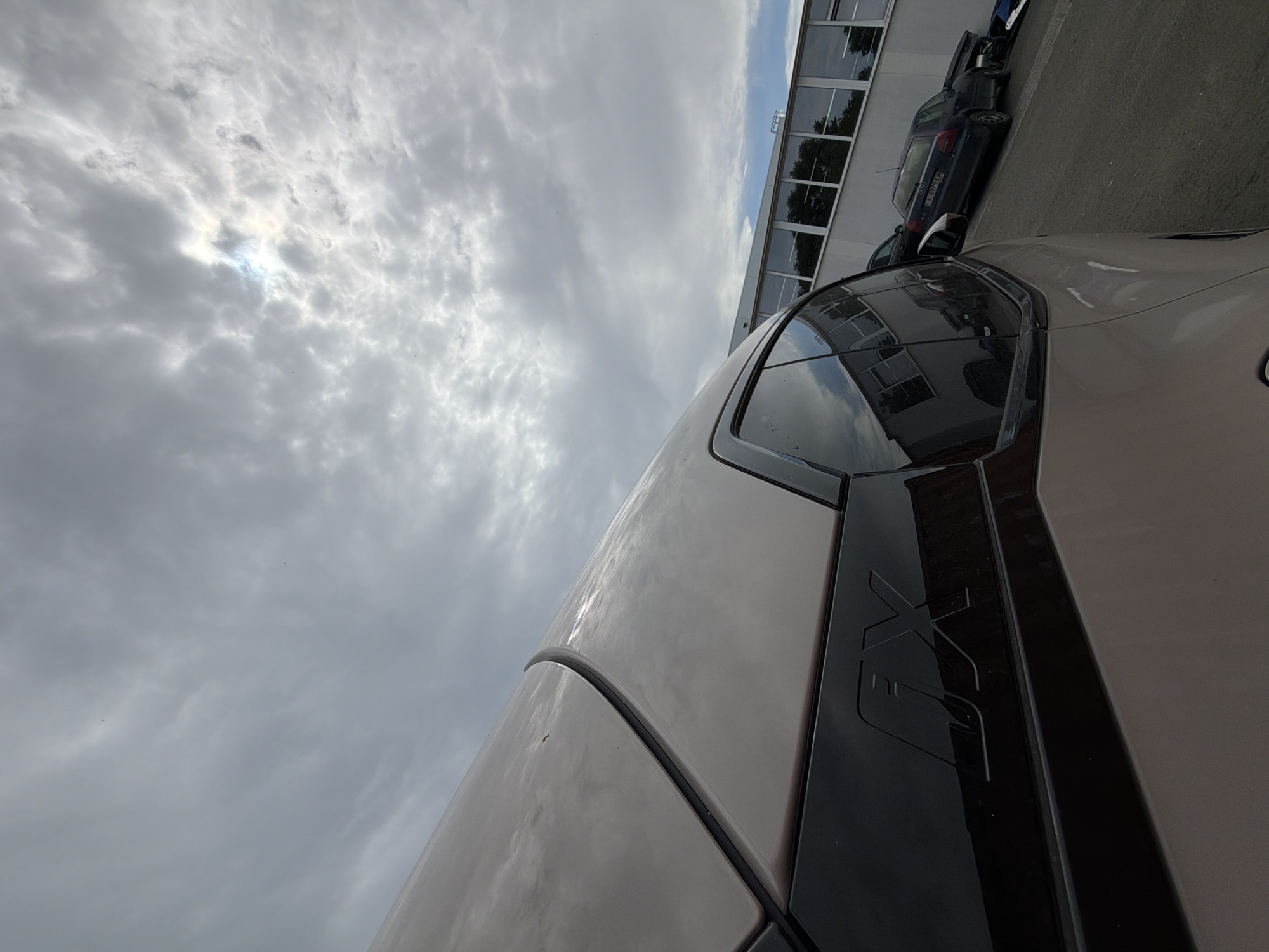 This media file shows a low-angle perspective of a modern vehicle, possibly a car, parked outdoors. The vehicle is light brown or beige and has a dark trim running along its side. A prominent, embossed logo with the letters "FX" is visible on the dark trim. The vehicle is angled in the frame, with the sky occupying a significant portion of the image.
The sky is overcast with scattered clouds, and the sun is visible through a break in the clouds, creating a bright, hazy effect. The lighting suggests it is daytime, likely mid-morning or afternoon. In the background, a building with large glass windows is visible, reflecting the sky and some trees. There are other vehicles parked behind the primary subject, including a dark-colored sedan with a license plate that appears to have Bulgarian characters.
The setting appears to be an urban or semi-urban environment, possibly a parking lot or an area adjacent to a commercial building. The overall mood is neutral, with no specific emotions conveyed. The image is oriented vertically, suggesting it was taken with the camera held in a portrait orientation. The presence of Bulgarian license plates on one of the vehicles indicates the location is Burgas, Bulgaria.