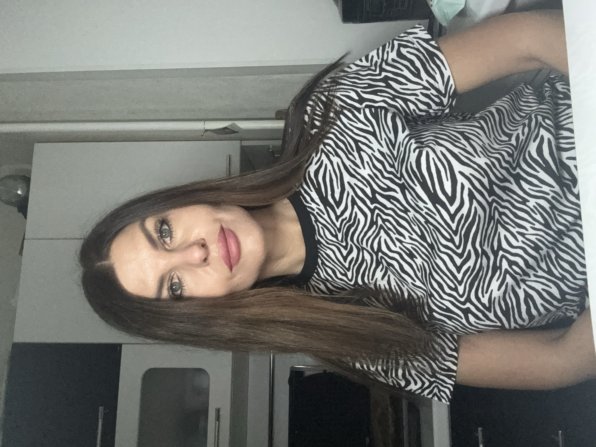 A woman with light skin, long brown hair, and light-colored eyes is wearing a black and white zebra-striped top. The background appears to be an indoor setting, likely a kitchen, with light gray cabinets and a silver fixture visible. The image is oriented sideways. There is no explicit evidence linking this scene to the city of Chaikovsky, Russia.