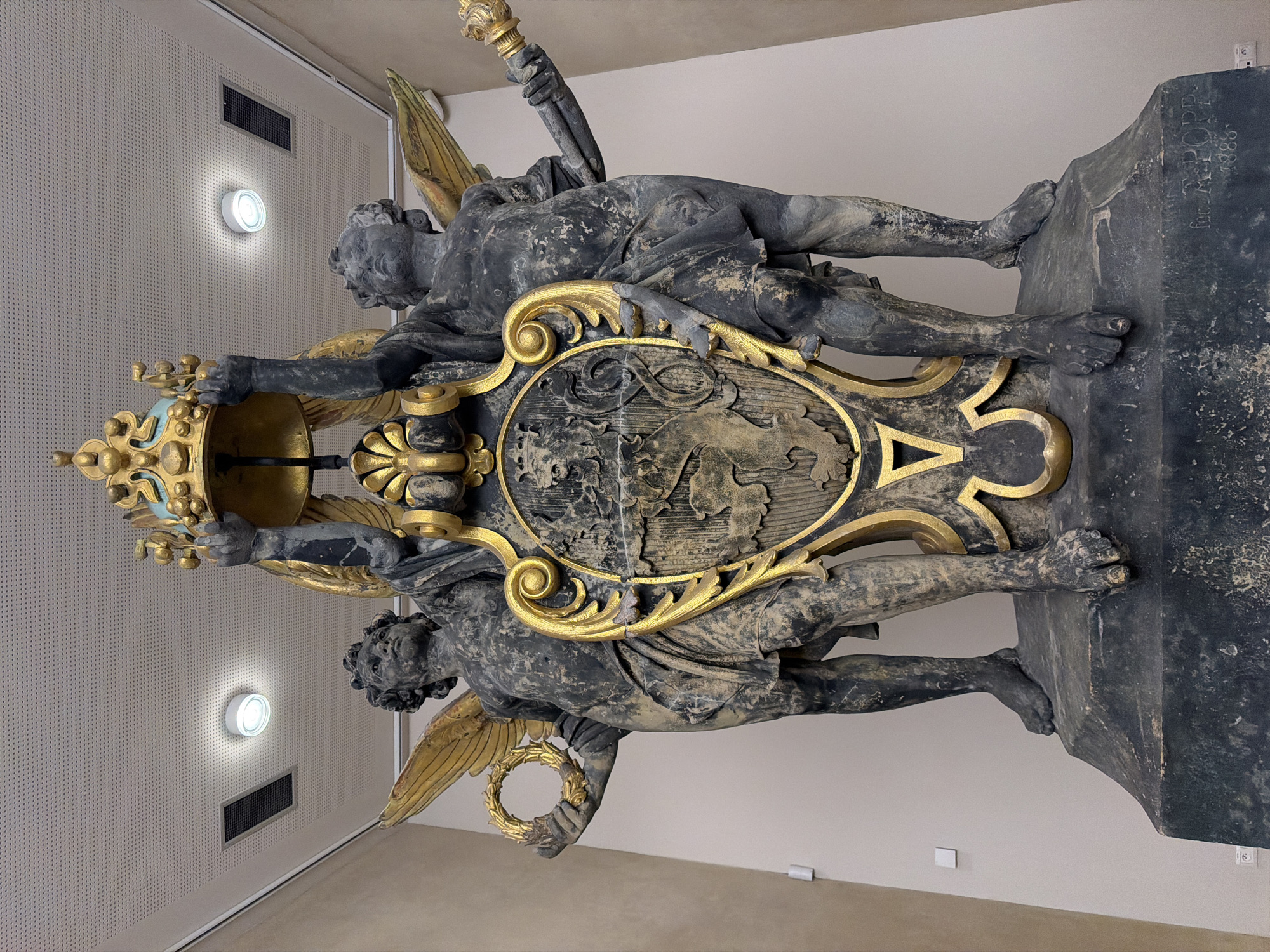 This is a low-angle, full shot of a dark stone sculpture depicting two winged cherubic figures holding up a golden crown and a shield. The figures are positioned on either side of the shield, with their arms raised to support the crown. The left figure holds a golden wreath in its left hand, and the right figure holds a golden scepter in its right hand. The shield itself is ornate and features a detailed coat of arms. The sculpture is placed on a dark stone pedestal, which has the inscription "For. A. POPP. 1888" carved into it. The background consists of a neutral-colored wall and a textured ceiling with lighting fixtures. The overall impression is one of grandeur and historical significance. The sculpture is likely located in an interior space, possibly a museum or public building. The style of the sculpture suggests it is from the late 19th century, consistent with the inscription on the pedestal.