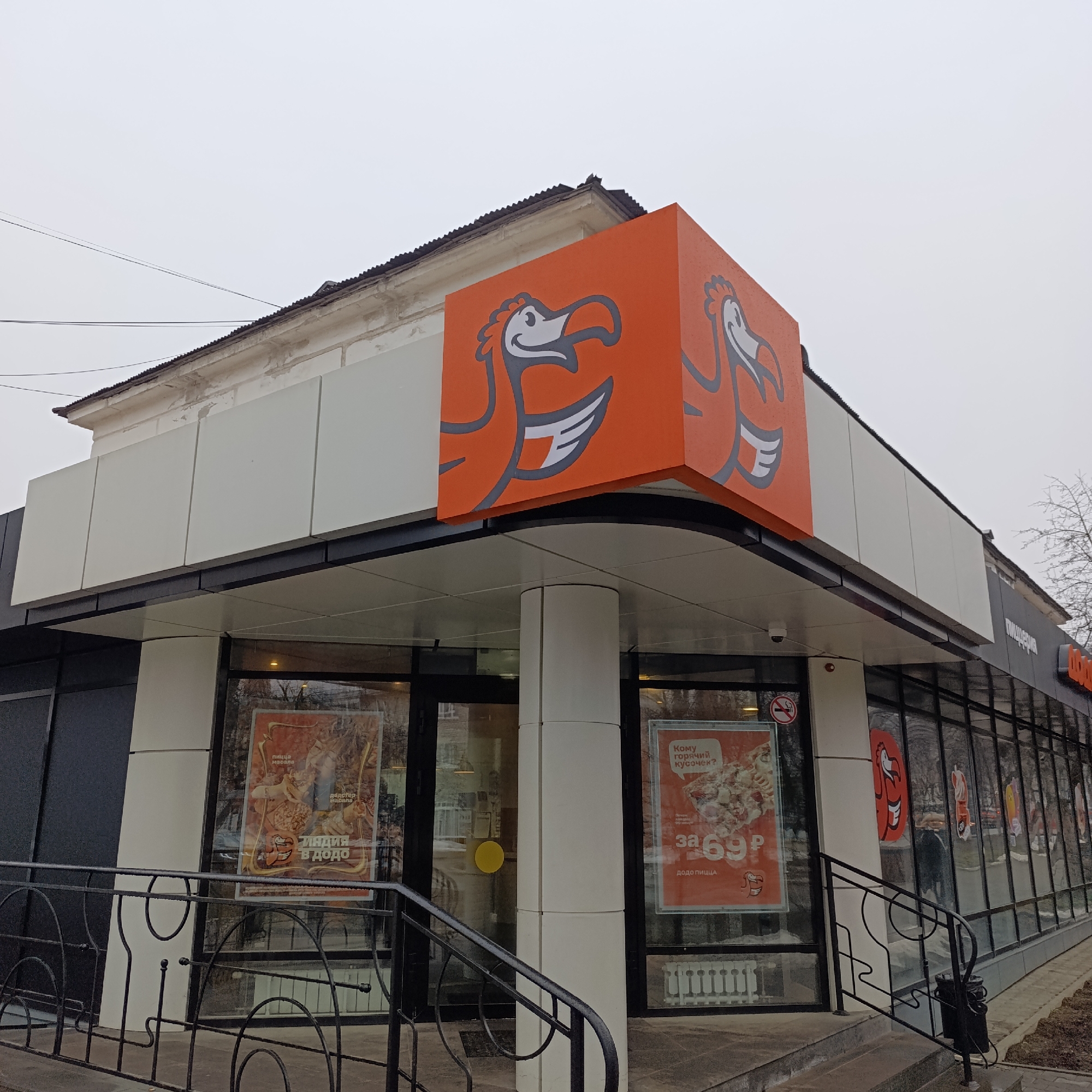 This is an eye-level outdoor shot of a Dodo Pizza restaurant in the city of Chaikovsky, Russia. The building has a modern design with a black and white exterior, featuring a large orange cube with the Dodo Pizza logo on top of the entrance. Posters are visible inside the glass windows. The outside environment is cloudy and urban, with a black metal railing leading up to the entrance.