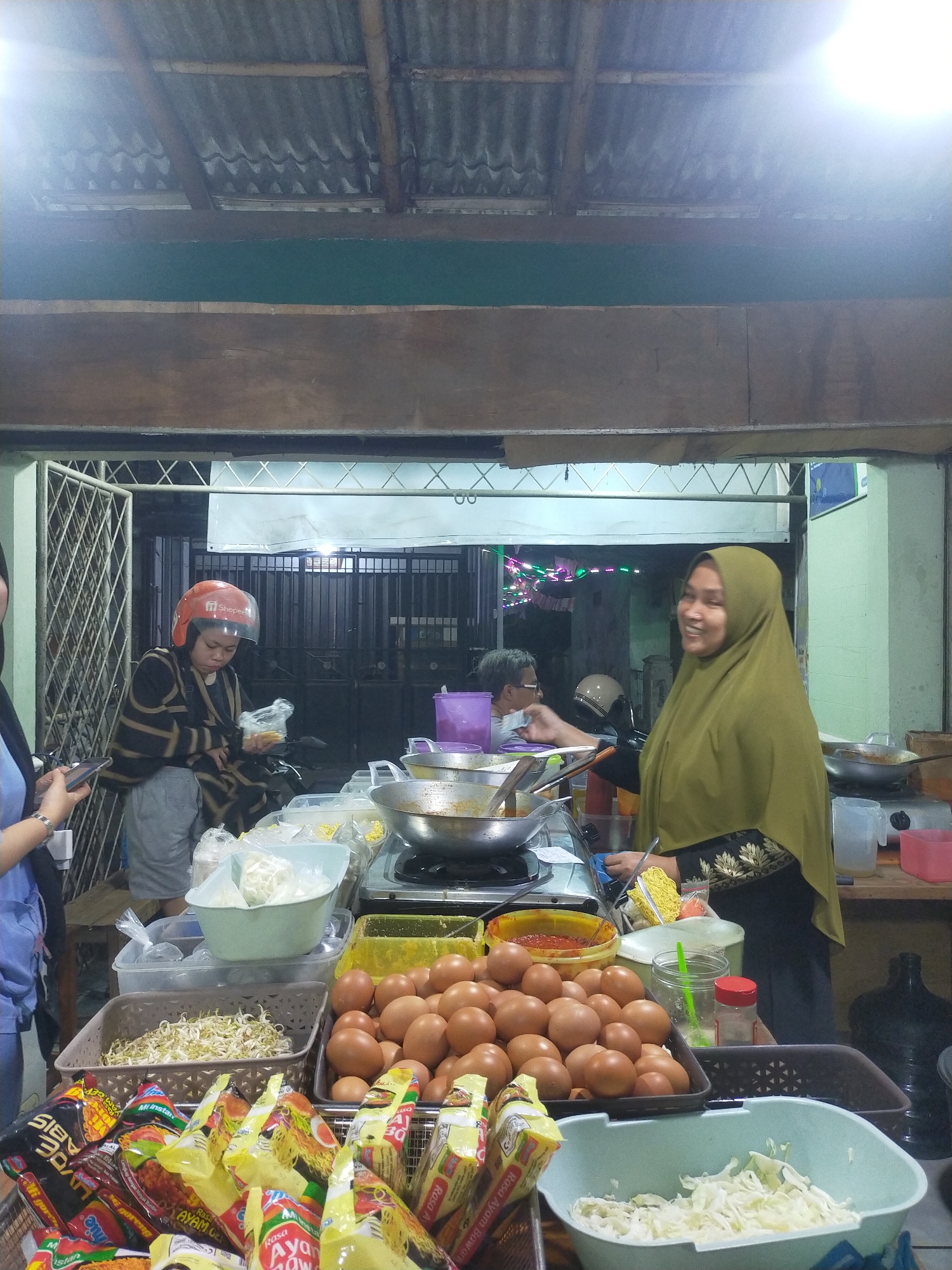 The image depicts an outdoor food stall at night, likely in an urban setting, given the presence of streetlights and the general atmosphere. The stall is set up under a corrugated metal roof supported by wooden beams. In the foreground, a variety of food ingredients and packaged goods are displayed. These include several packages of instant noodles, a large quantity of raw eggs, a basket of bean sprouts, a bowl of shredded cabbage, and a bowl of what appears to be chili paste.
Two women are the primary subjects. On the right, a woman wearing a green hijab and a black garment with gold embroidery is smiling and appears to be interacting with someone just out of frame. She is standing behind the food stall, possibly as the vendor. On the left, another woman, partially visible, is wearing a blue top and holding a smartphone. In the mid-ground, a woman wearing a black and yellow striped jacket and a red motorcycle helmet is holding a plastic bag containing food. She appears to be a customer or another worker at the stall. Behind her, another person, likely a customer, is seated and wearing glasses.
The stall itself has cooking equipment, including a large metal wok on a stove, suggesting food is being prepared. Various containers and utensils are visible. The background shows a dark metallic gate or fence, possibly leading into a building or another area, and further back, there are decorative string lights, suggesting a festive or active evening environment. The overall scene is characteristic of a local street food vendor, with a focus on fresh ingredients and customer service.