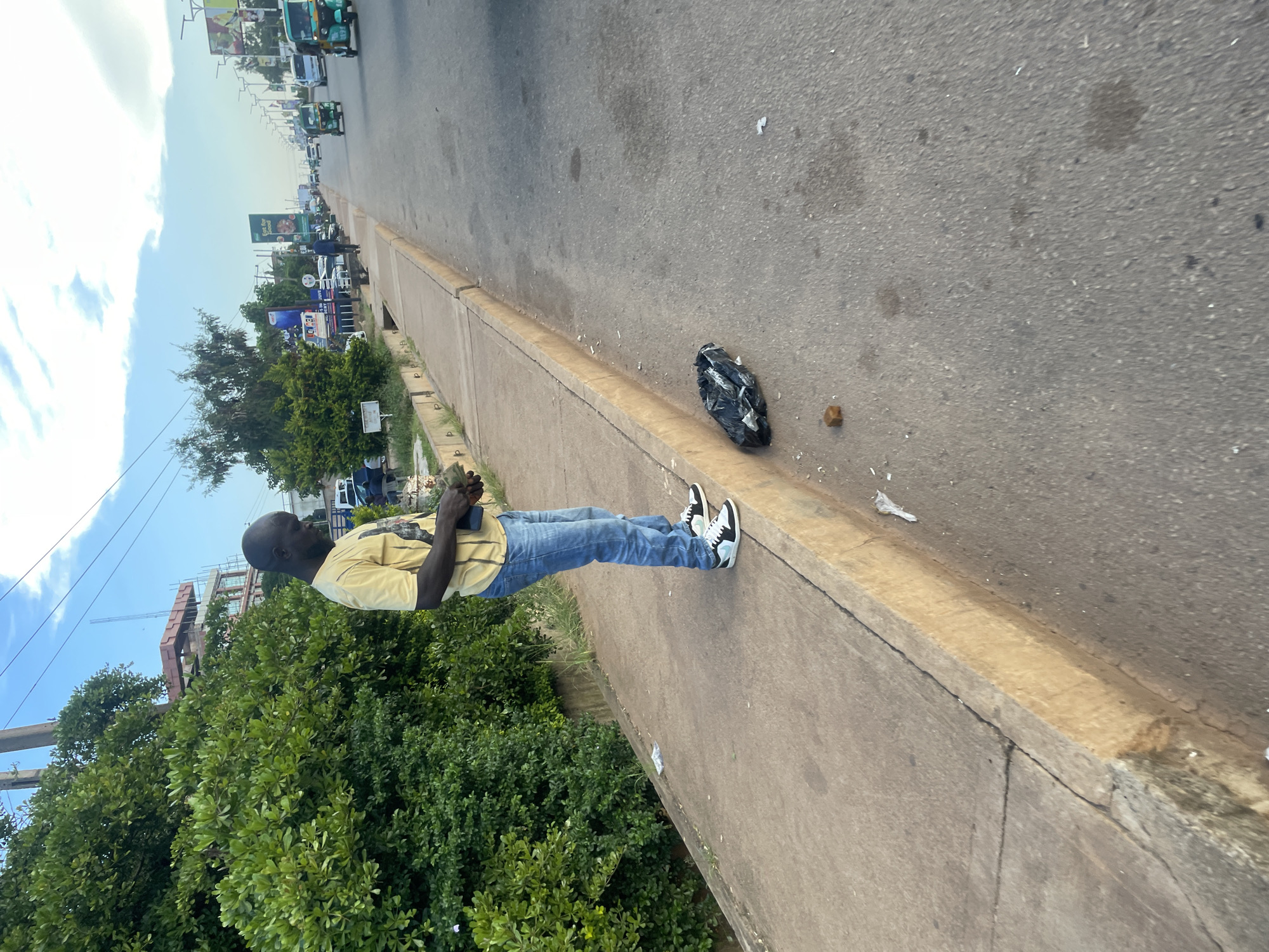 A man stands on a sidewalk in Jos, Nigeria, holding a wad of cash in his hands. He is wearing a yellow t-shirt and blue jeans, and he has a shaved head. A black trash bag lies on the road next to the sidewalk, and a small brown object, possibly a brick, is on the sidewalk nearby. The background shows a street with traffic, including cars and a few three-wheeled vehicles, and several buildings and billboards. The sky is blue with some clouds.