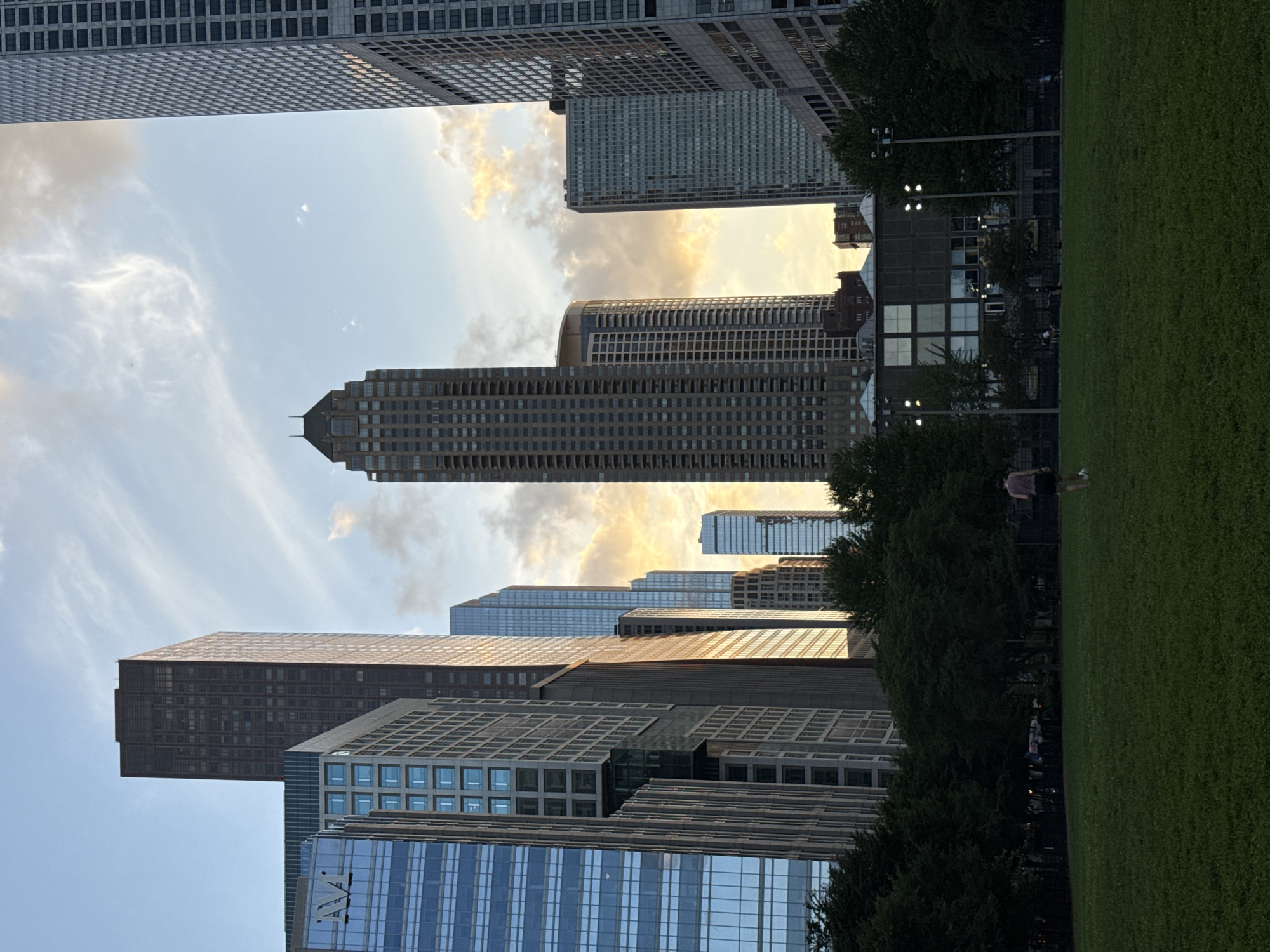 The image depicts a park in Chicago, Illinois, with a backdrop of towering skyscrapers. The park features a large expanse of manicured green lawn in the foreground. A lone person is walking on the lawn. Beyond the lawn, a line of mature trees separates the park from a dark building with a patterned facade and illuminated windows. Further beyond the trees, the city skyline rises, dominated by numerous high-rise buildings of varying architectural styles and heights. The sky above is a blend of blue and soft clouds, with hints of golden sunset light peeking through, suggesting the time of day is late afternoon or early evening. The overall scene conveys a sense of urban tranquility juxtaposed with the impressive scale of the city's architecture.