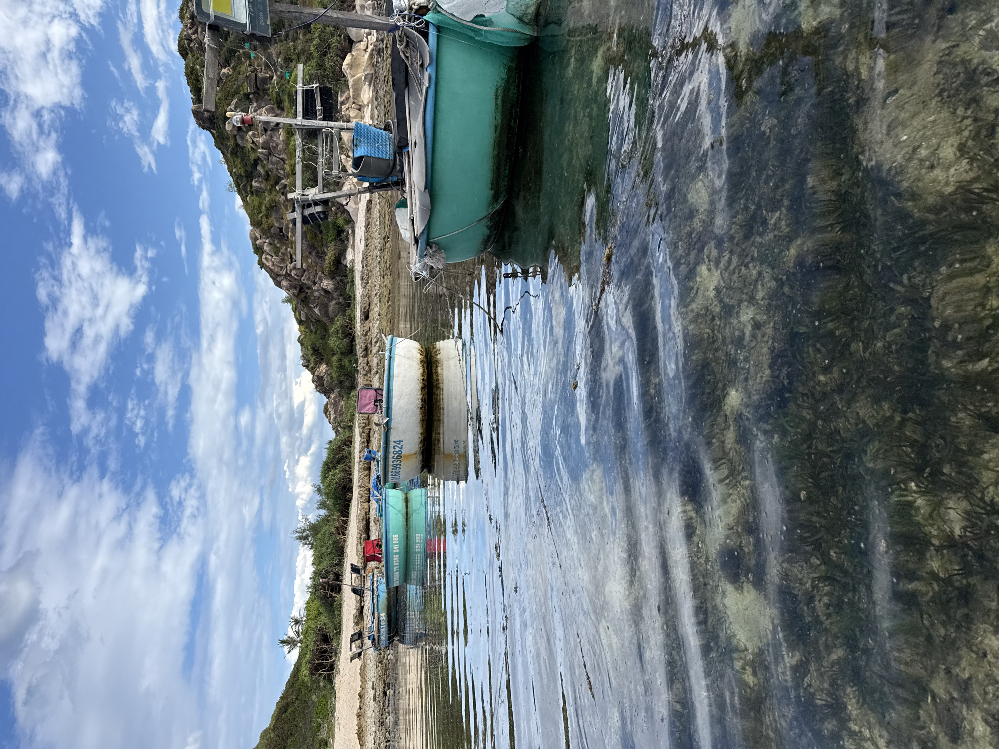 In the coastal area of Từ Nham, Vietnam, the image captures a tranquil scene with several small fishing boats anchored in shallow, clear water. The boats are of traditional design, with rounded hulls, and are positioned close to a sandy shore lined with sparse vegetation. Some boats are brightly colored, featuring green and white hulls, while others are a muted blue. Visible details on the boats include wooden seating, fishing equipment, and weathered hulls suggesting recent use. The seabed beneath the water is visible, showing patches of seaweed and sand, indicating a calm and shallow environment. In the background, a rocky, vegetated hillside rises, with some evidence of human presence such as poles and wires. The sky above is a vibrant blue, dotted with scattered white clouds, contributing to the overall serene and picturesque atmosphere of the location.