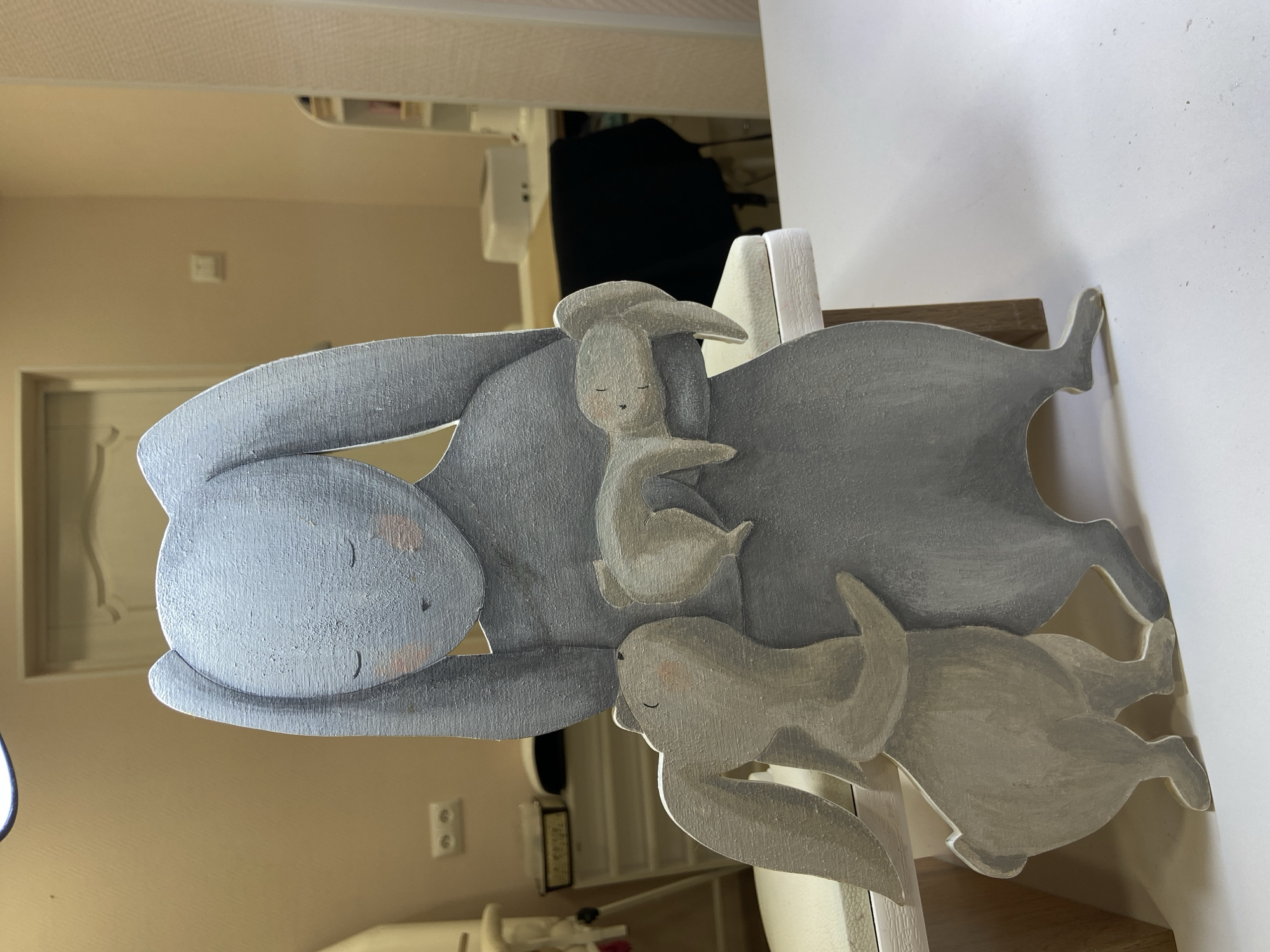A painted wooden sculpture of a mother rabbit with two baby rabbits, one on her back and one at her feet, is depicted. The sculpture is painted in shades of gray. It sits atop a white shelf or piece of furniture, which itself rests on a wooden surface. The background shows a room interior with beige walls, a door, and other furniture partially visible. There is no indication that this image was taken in Chaikovsky, Russia.