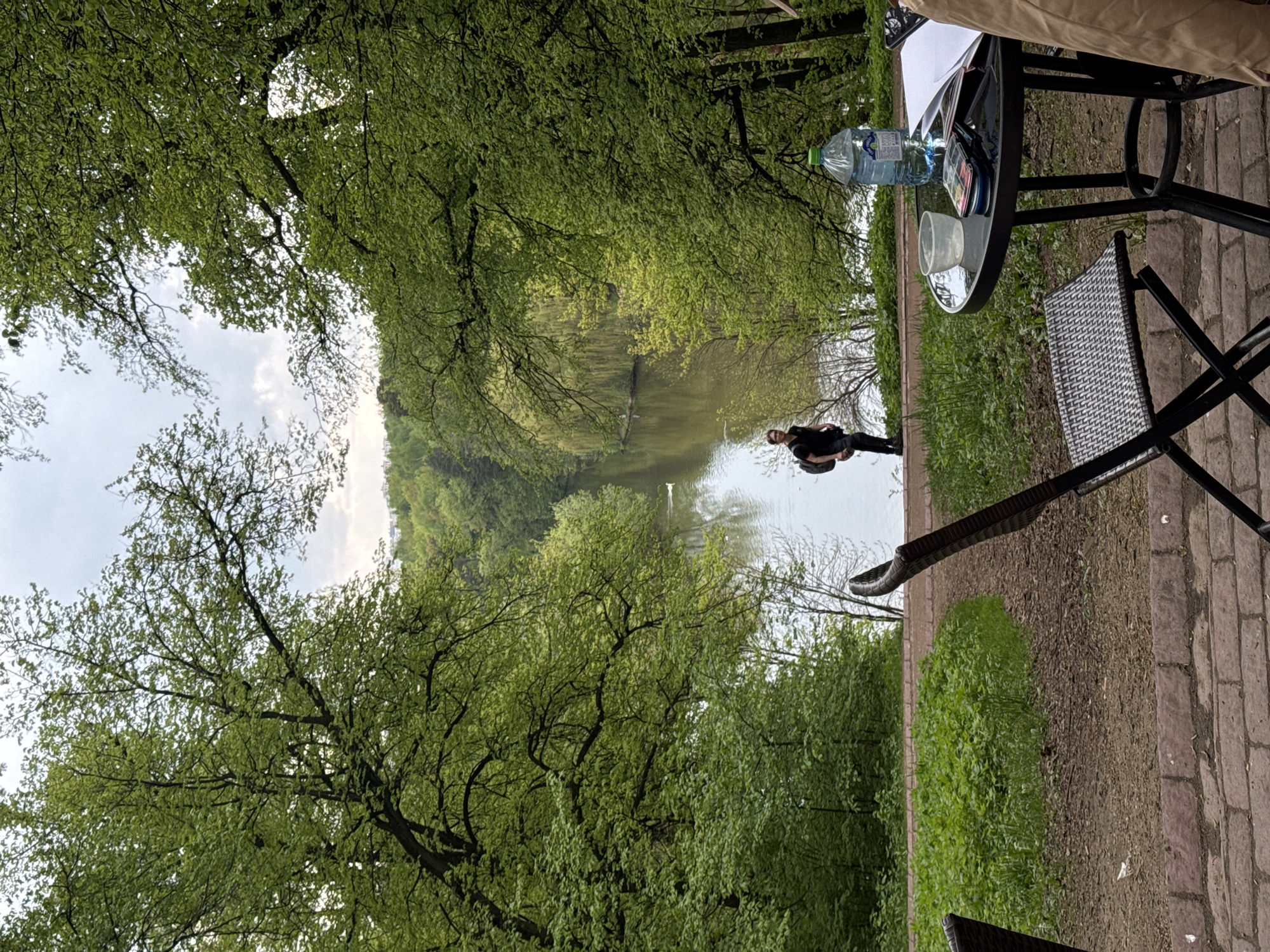 The image shows a landscape view of a park in Moscow, Russia, featuring a long pond or narrow lake surrounded by lush green trees. A person wearing dark clothing stands near the water's edge. A table with a glass top and a black metal frame is visible in the foreground, along with a folding chair with a patterned seat and back. A water bottle, a glass, and what appears to be an artist's palette are on the table. The ground in the foreground is brick, and there is greenery visible between the bricked area and the water. The sky is overcast with light clouds.