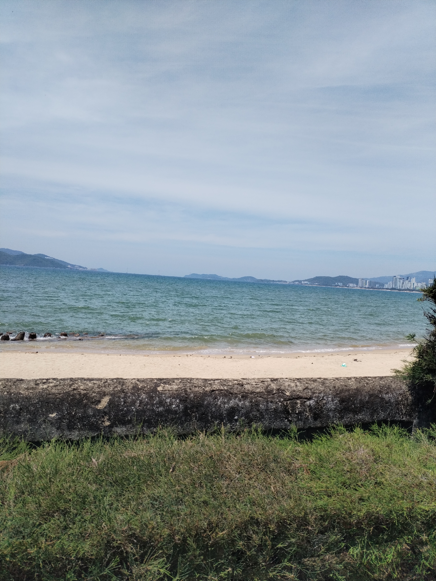 This is a photograph of a beach in Nha Trang, Vietnam. The sky is overcast with light clouds. The ocean is a deep blue-green with moderate waves. The beach is sandy and appears to be mostly empty, with a few scattered pieces of debris. In the foreground, there is a low, gray concrete wall, and behind that, dense green grass. In the distance, across the water, there is a line of hills and a city skyline with several tall buildings. The water appears relatively calm, and the overall atmosphere is serene and somewhat hazy.