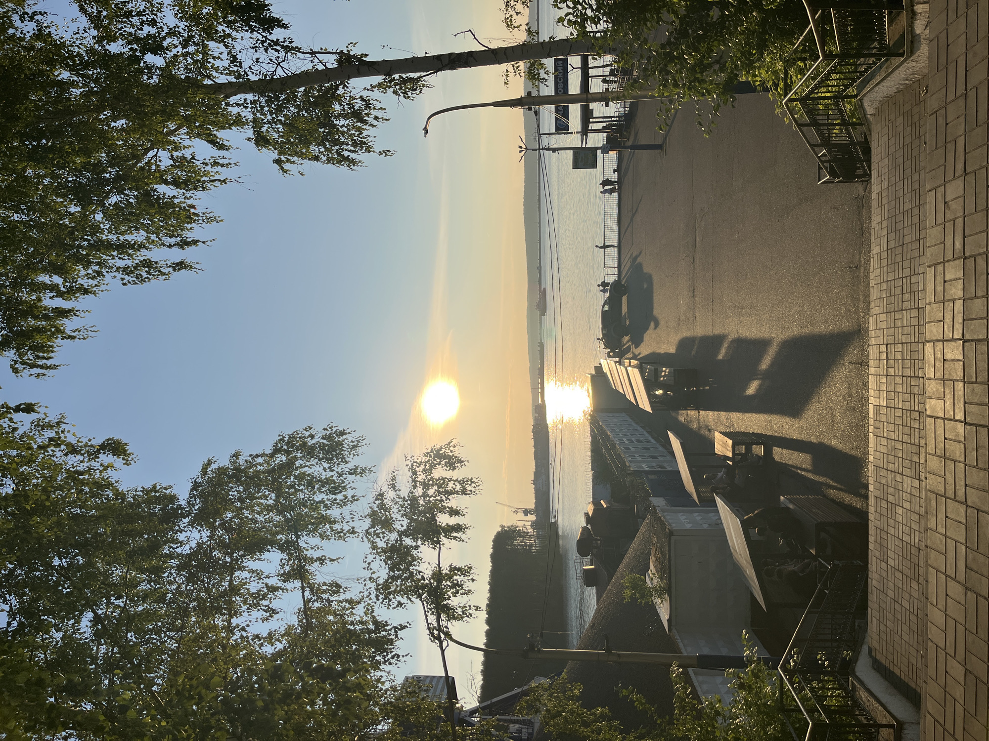 This image depicts an outdoor scene at sunset, likely in Chaikovsky, Russia. The sun, a bright orb, is low in the sky, casting a warm, golden light and reflecting brilliantly on the surface of a body of water, possibly a river or reservoir. The scene is framed by lush green trees with leaves that appear to be rustling, suggesting a gentle breeze.
In the foreground, a paved pathway runs alongside the water, bordered by a metal railing. The pathway is lined with several benches, some of which are occupied by silhouetted figures of people. Beyond the pathway, there are some structures that appear to be small buildings or kiosks. Further out on the water, a ship or boat is visible, adding to the sense of a working waterfront or recreational area. The sky is a clear blue, fading to a soft yellow and orange near the horizon where the sun is setting.
A lamppost with a bent pole stands near the pathway, casting a long shadow. There is also a parked vehicle, possibly a scooter or small car, on the paved area, its shadow also stretching across the ground. The overall atmosphere is peaceful and serene, capturing the beauty of a summer evening by the water. The textured paving stones of a walkway are visible on the right side of the image. A sign with Cyrillic text is partially visible on a post near the water's edge.