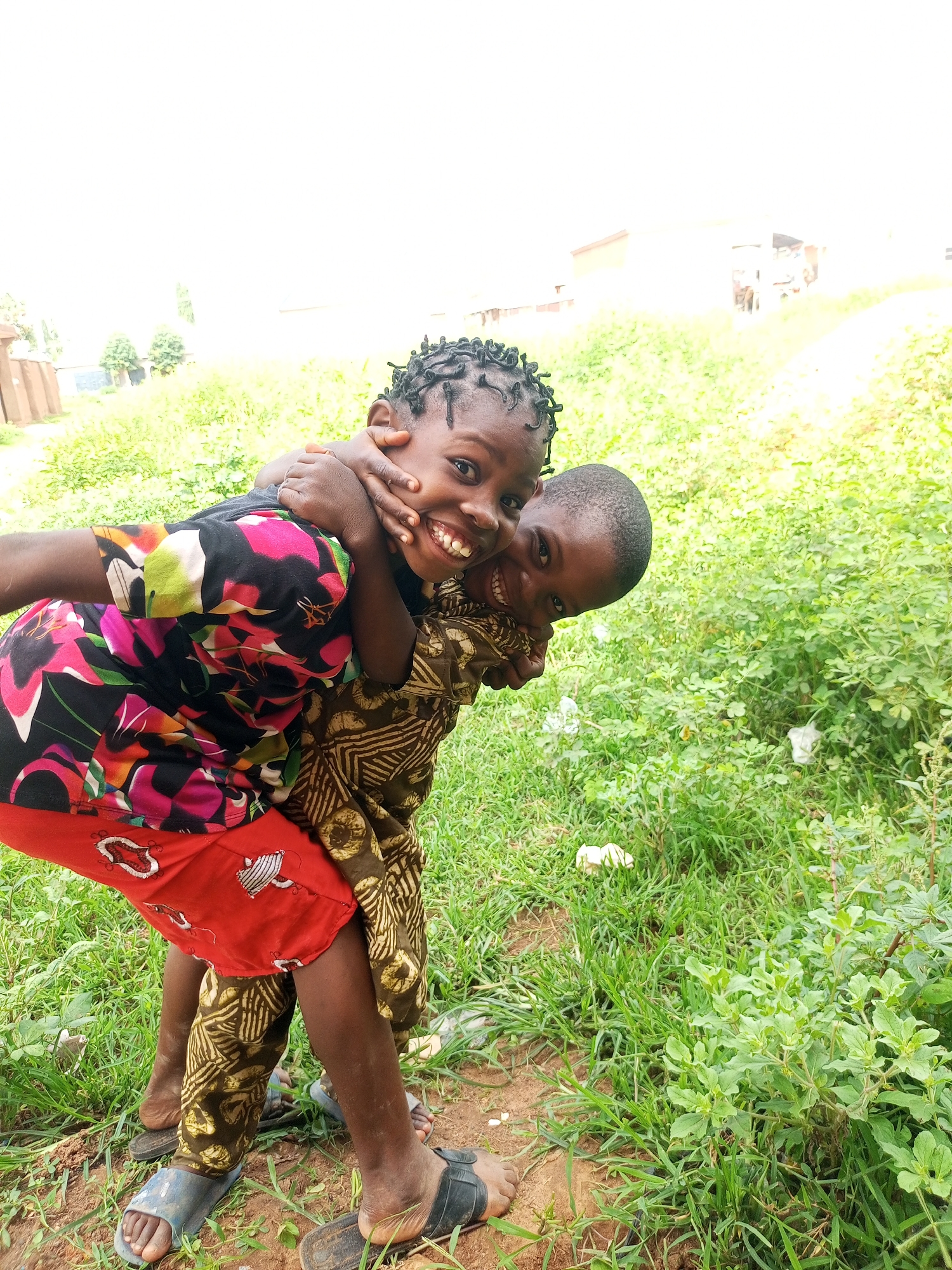 This is a close-up, waist-up portrait of two young children, a boy and a girl, in what appears to be an outdoor setting in Gombe, Nigeria. The girl, positioned in the foreground and on the left, is being piggybacked by the boy. She is looking towards the camera with a wide, joyful smile, her arms wrapped around the boy's neck. Her hair is styled in numerous small braids, and she is wearing a black t-shirt with a colorful floral print in shades of pink, yellow, and green, along with a vibrant red skirt or shorts with a white pattern. The boy, behind her, is also smiling, his head tilted and looking slightly off-camera. He is wearing a patterned brown garment. They are both wearing casual footwear, with the girl's feet and sandals visible at the bottom of the frame. The background consists of lush green grass and foliage, with some dried leaves and possibly some litter visible. The overall impression is one of playfulness and happiness.