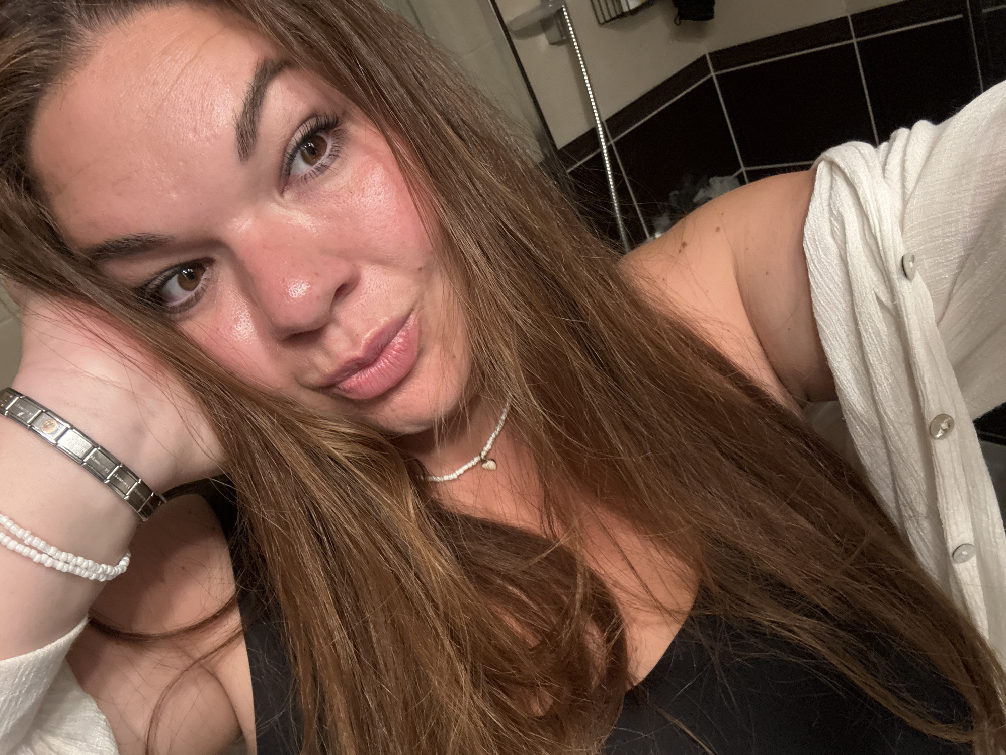 A woman poses for a selfie indoors. She is fair-skinned, with brown eyes and long brown hair. She is wearing a black tank top and a white cardigan or shirt. On her left wrist, she has a metal bracelet and a beaded bracelet. She is also wearing a beaded necklace with a small charm. The background includes black and white tiles, potentially in a bathroom setting. Based on the prompt, the location is Bijela, Montenegro, but there are no visible elements within the image that confirm this.