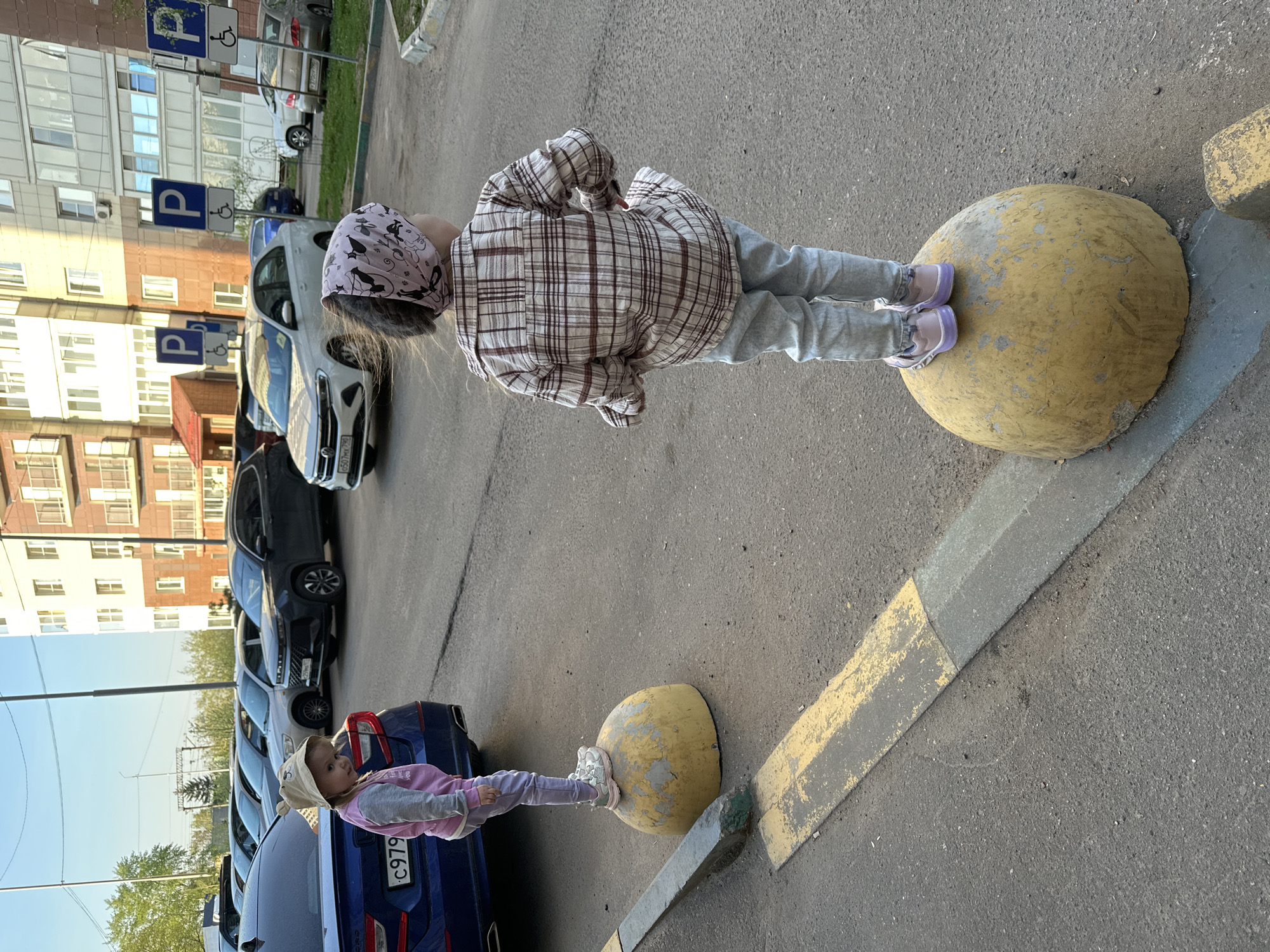 Two girls are standing on concrete spheres in what appears to be a parking lot. The girl on the right, who is taller, is wearing a plaid shirt, gray pants, and a head covering. The shorter girl is wearing a light pink and gray outfit and a sun hat. Cars are parked in the background, along with a building with handicap parking signs. The location is Tomilino, Russia.