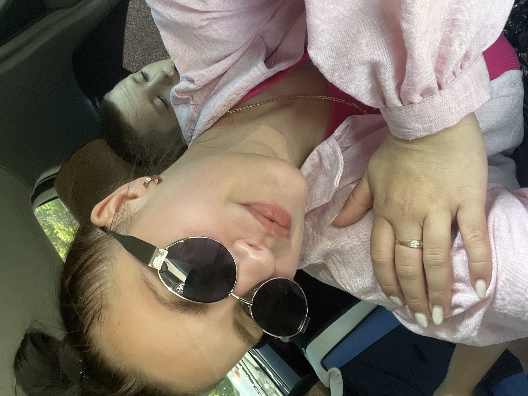 A woman with sunglasses and a pink shirt is in a car. There is a child asleep next to her. The location is unknown.