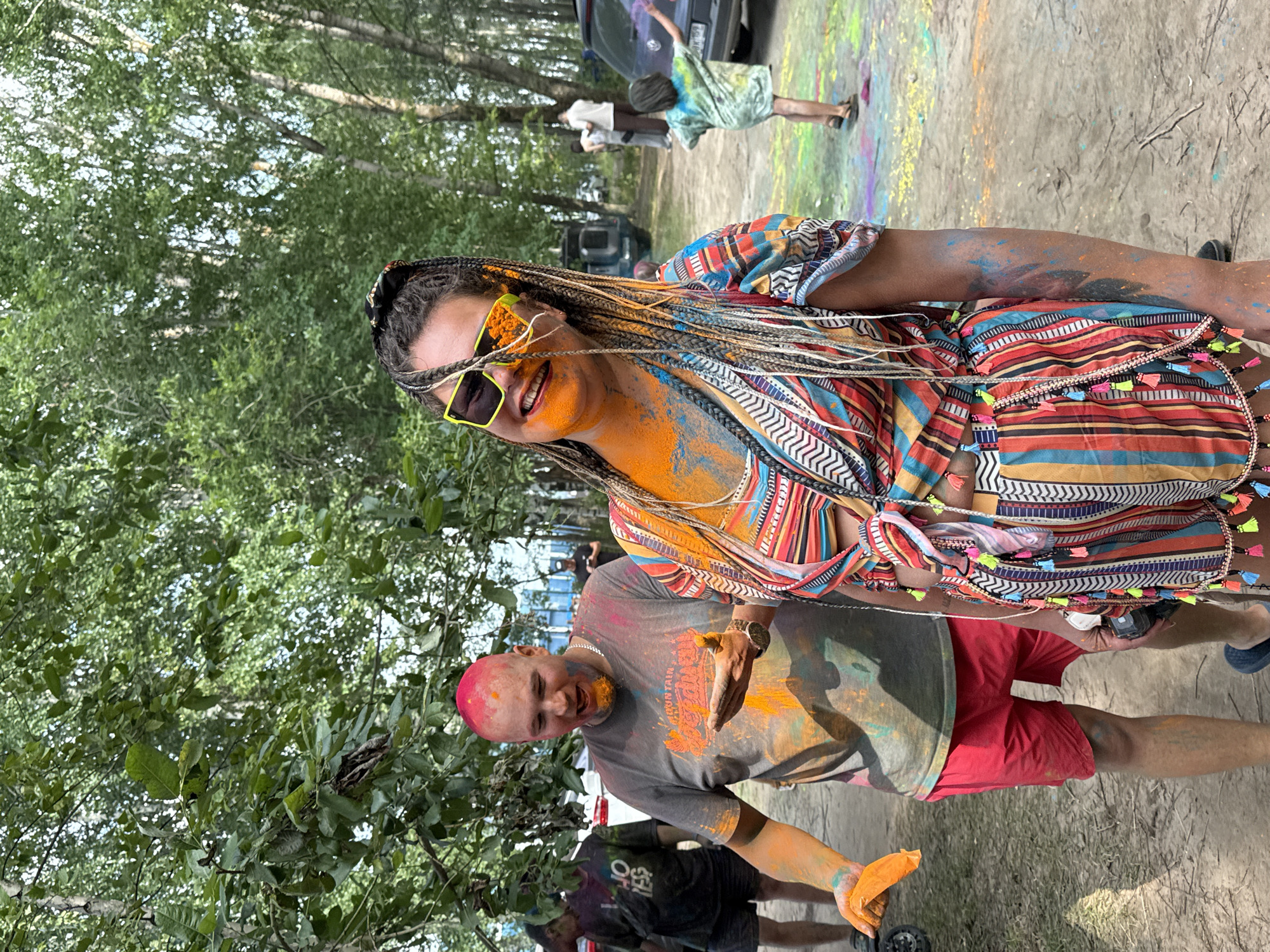 In a wooded area, a group of people are participating in a color powder festival. The main subject is a woman with her hair in braids, covered in orange and blue powder. She is wearing a colorful, patterned outfit and bright yellow sunglasses. To her left, a bald man with pink-colored hair is also covered in powder, holding a handful of orange powder. In the background, other individuals are visible, some with powder on them, and cars are parked in the distance. The ground is dirt and scattered with colorful powder.